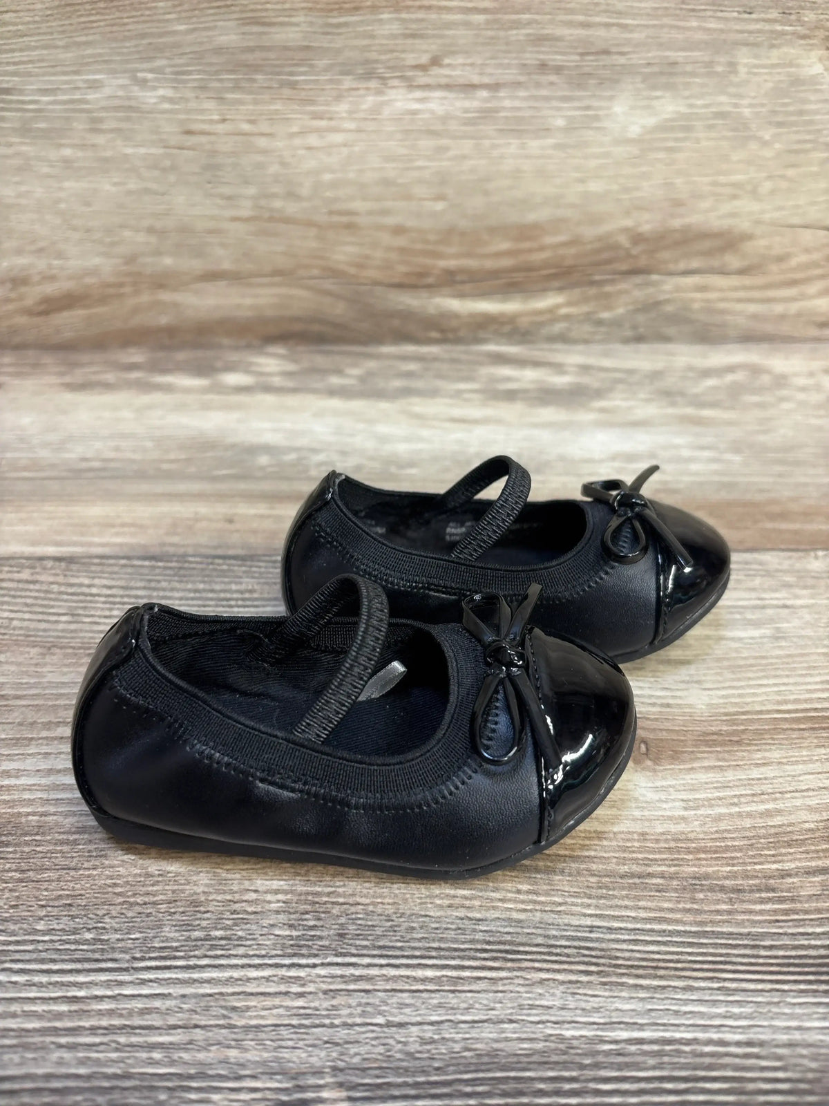 Angled side view of black toddler Mary Jane flats with elastic strap, patent toe and bow on wood background
