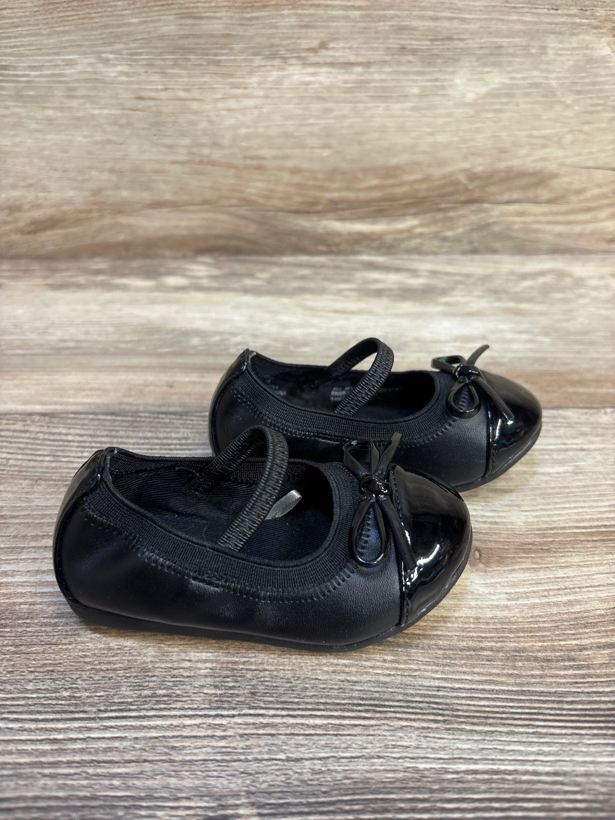 Angled side view of Children's Place Mary Jane flats size 4C in black with elastic strap, patent toe cap and bow on wood background