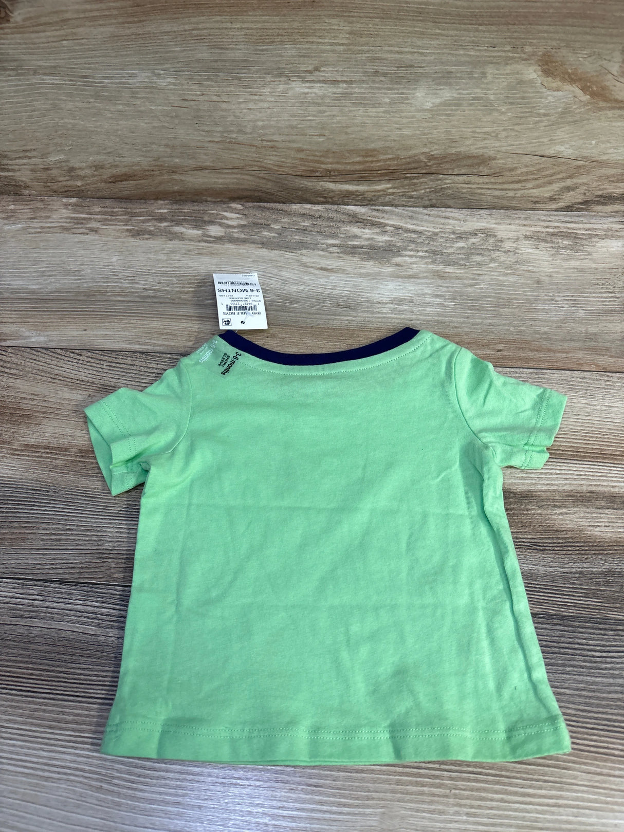 Back view of First Impressions skateboarding snail shirt in light green showing navy crew neckline, short sleeves, and size tag on wood floor