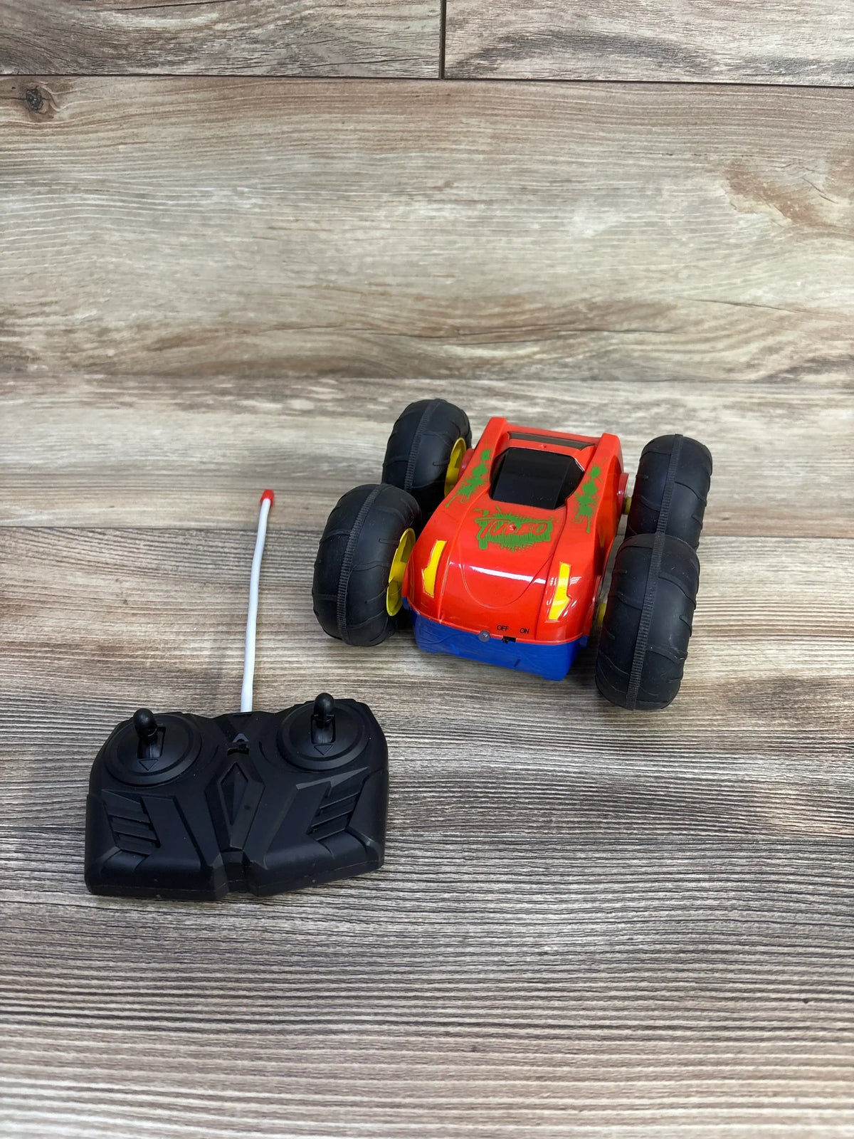 Top view of Turbo Wheel Radio Control Stunt Racer Car showing four large wheels, remote control, and double-sided design on wood-grain surface