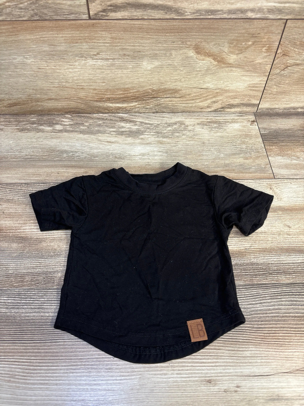 Front view of Little Bipsy black bamboo shirt in size 3 to 6 months, showing short sleeves and curved hem on wood-look floor