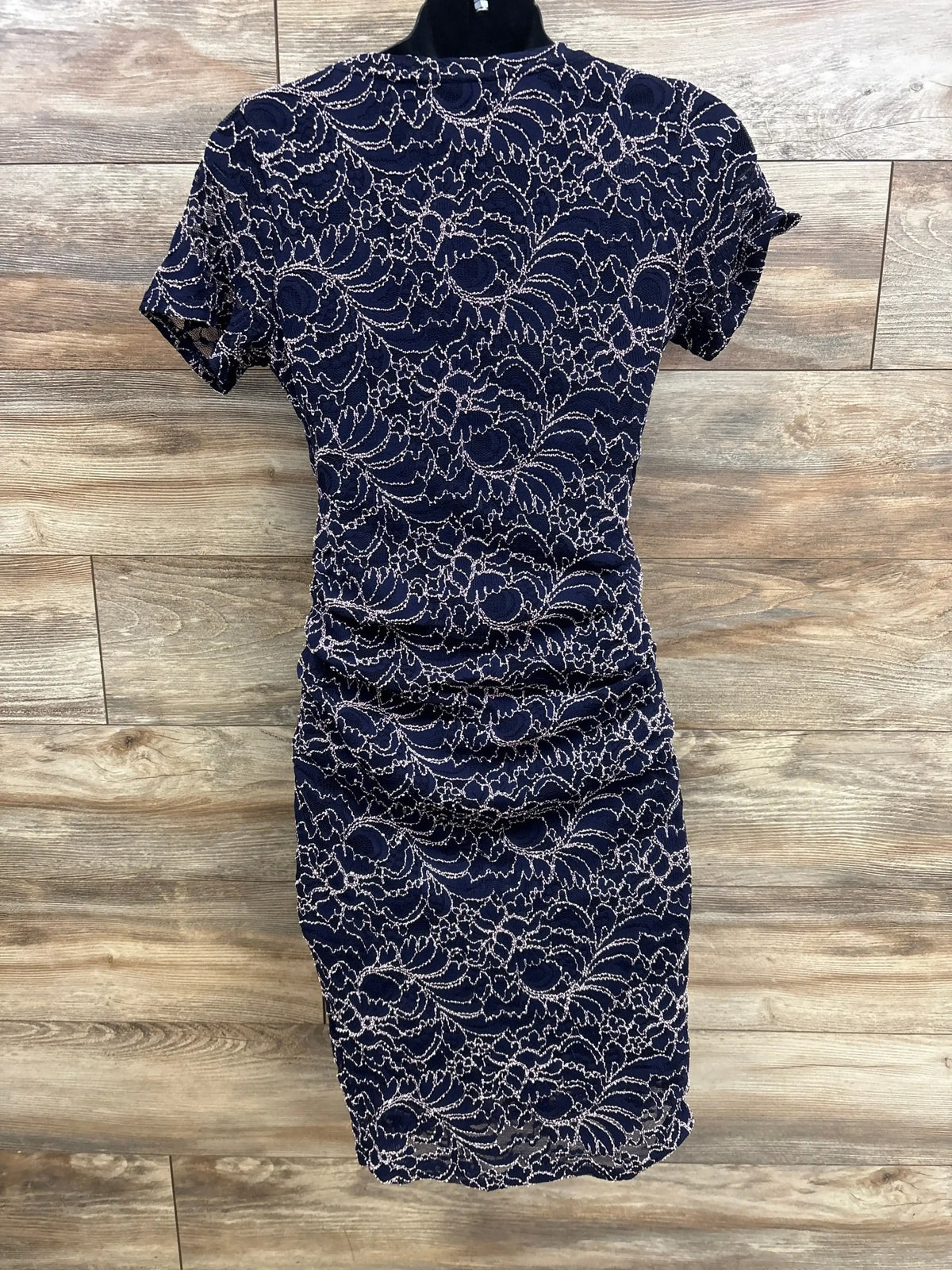 Front view of Motherhood Maternity navy lace maternity dress with short sleeves, ruched sides, and floral lace overlay on wood plank background