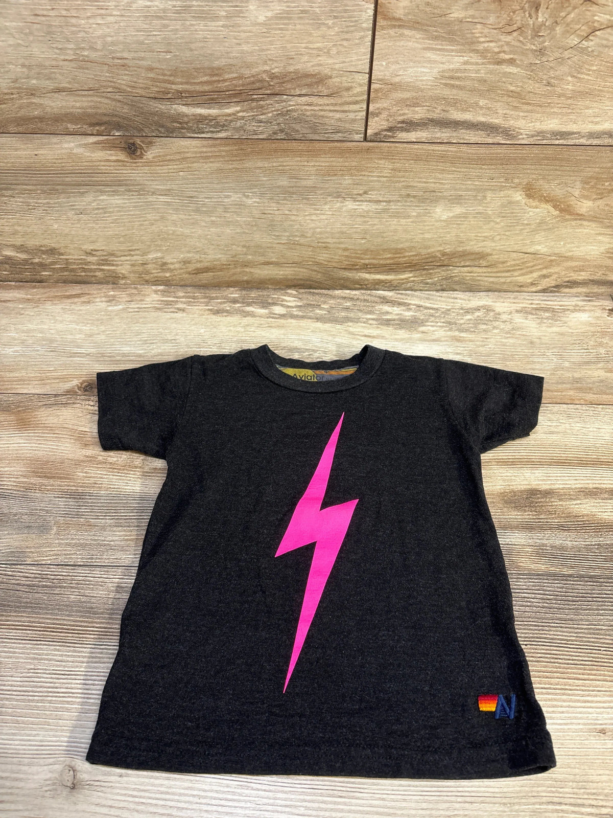 Flat lay of Aviator Nation 4T black toddler tee showcasing pink lightning bolt and small rainbow AN logo on light wood floor