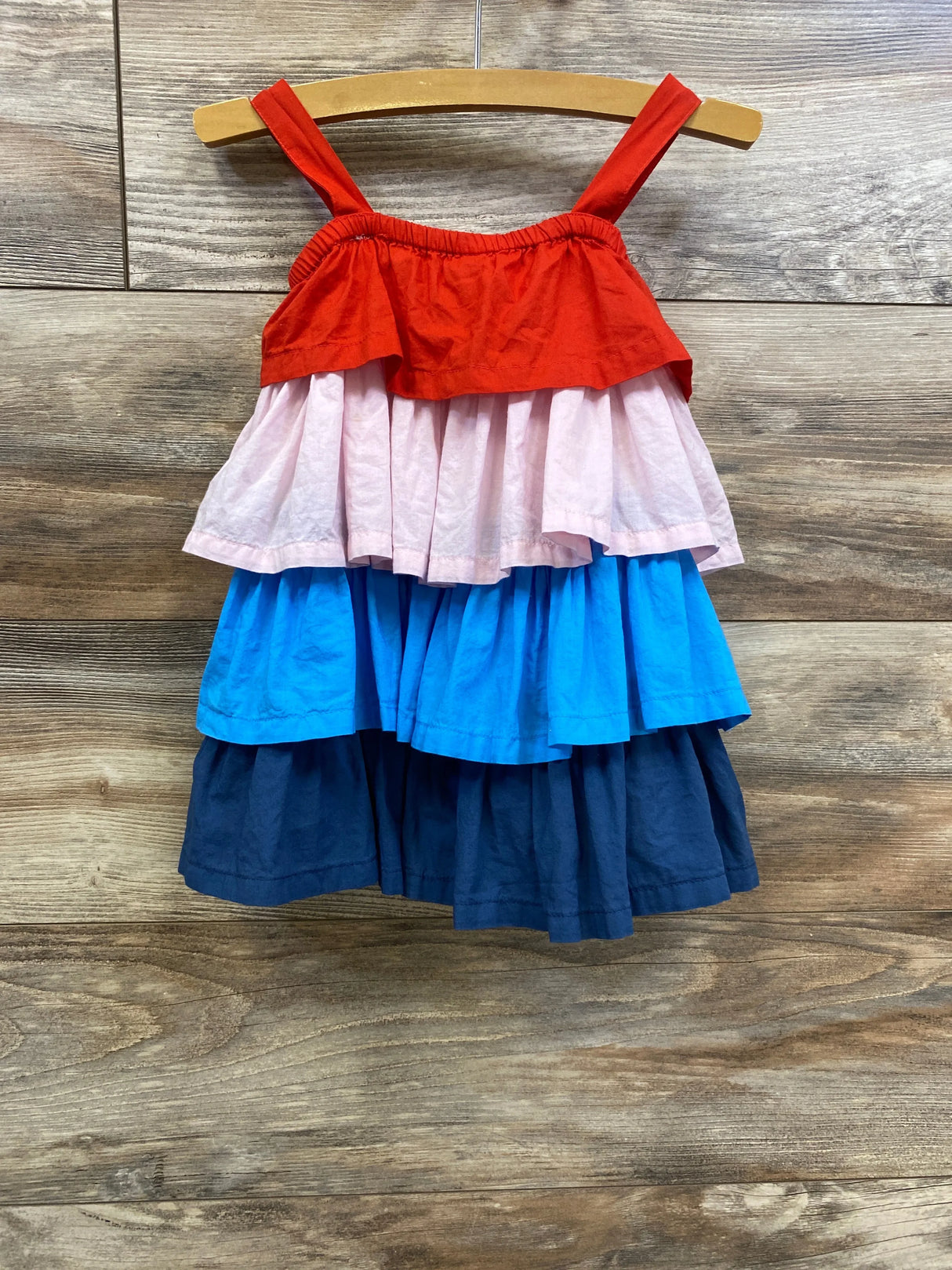 Front view of tiered toddler dress with red, pink, bright blue and navy tiers on wooden hanger against wood plank background