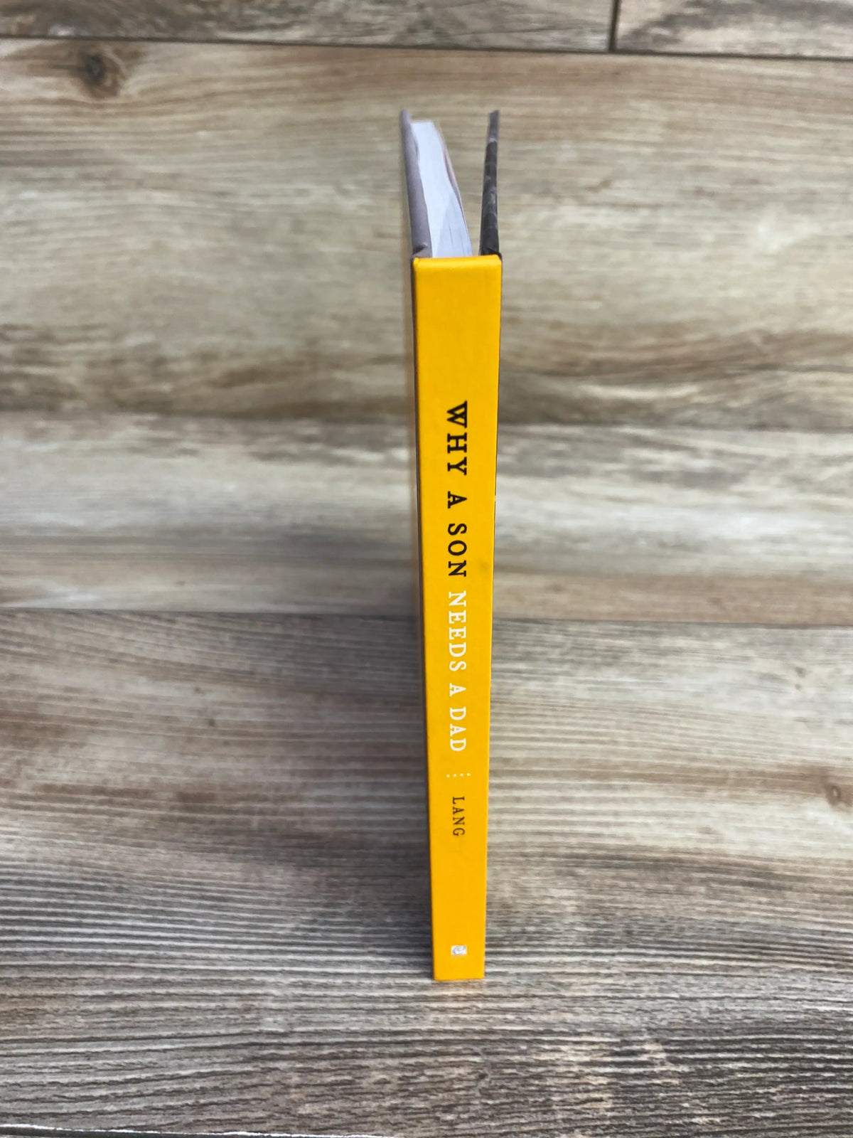 Spine view of Why a Son Needs a Dad hardcover book by Lang showcasing yellow spine with white text on wood plank floor
