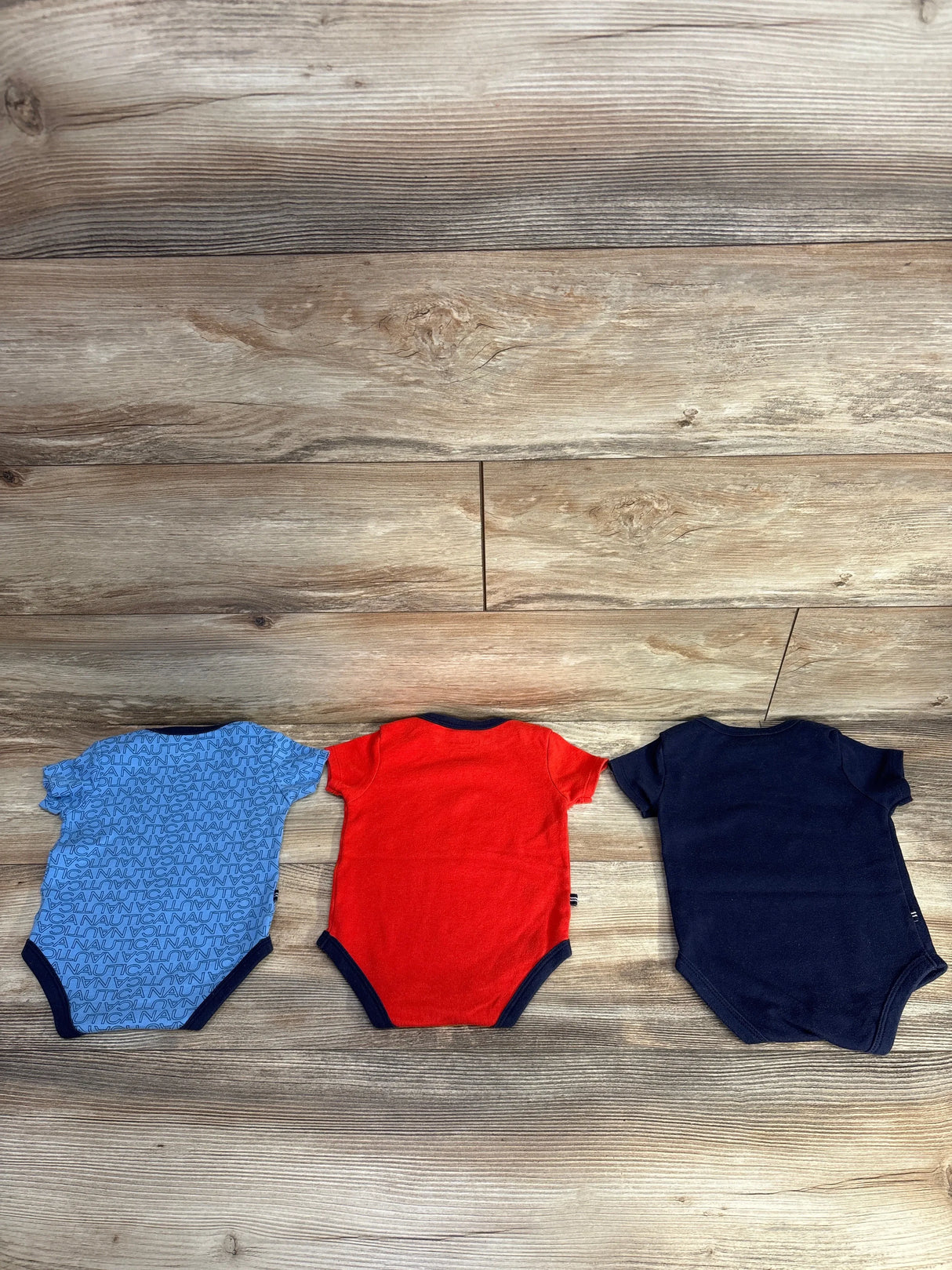 Three short-sleeve baby onesies in blue with Nautica text, red, and navy blue on wood floor
