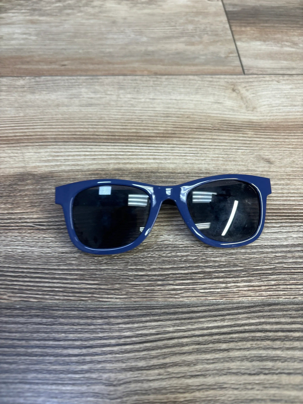 Front view of kids' blue plastic sunglasses showing rectangular frame and dark lenses on wood tabletop