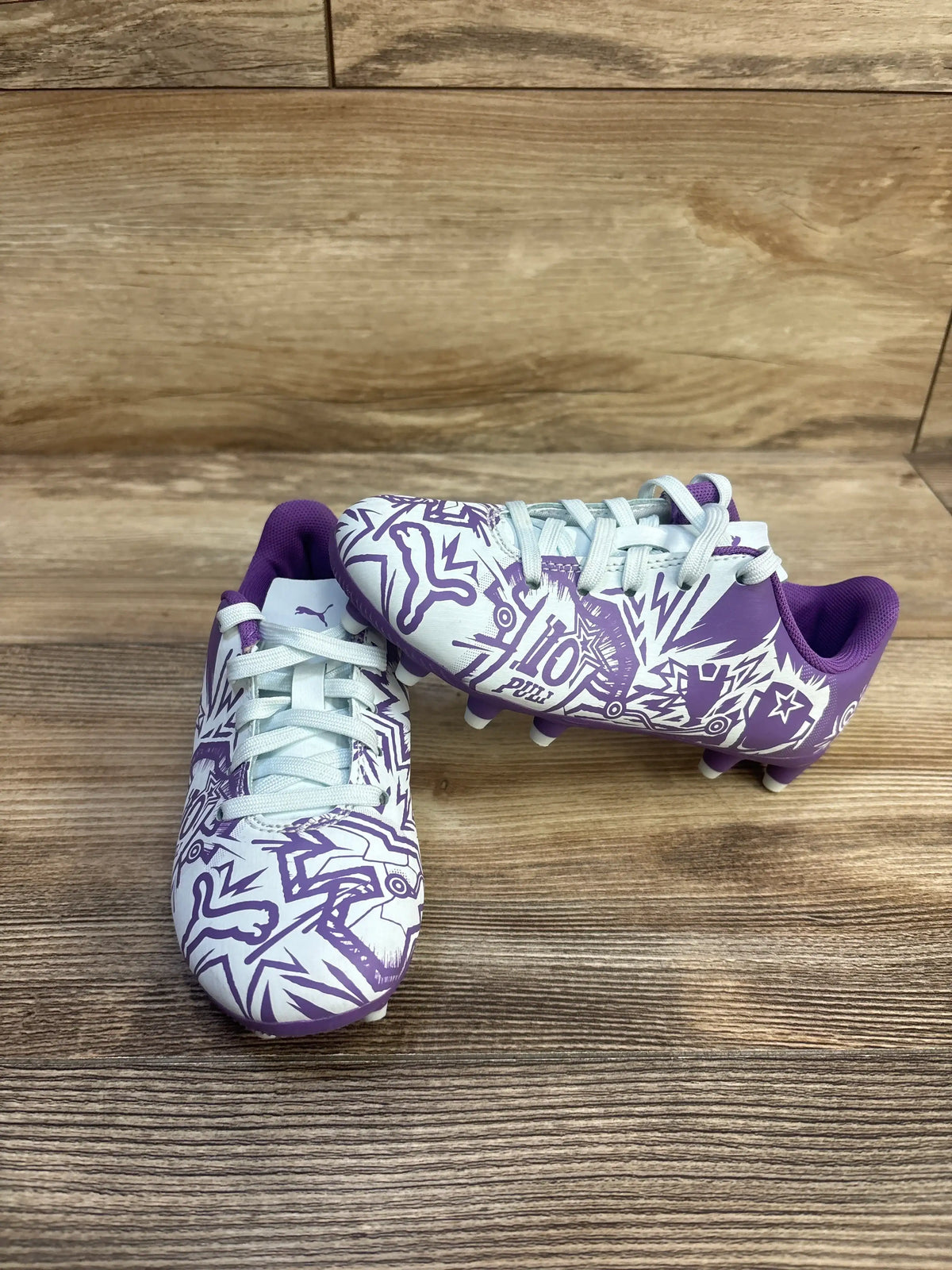 Side view of Puma x Christian Pulisic Tacto II soccer cleats showing purple-white graphic uppers and molded studs on wood floor