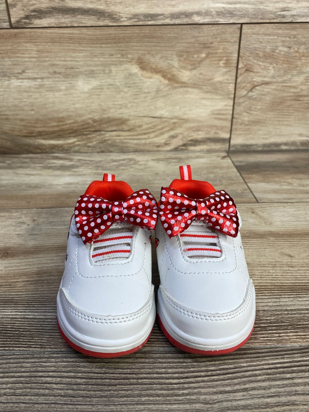 Front view of Disney Baby Minnie Mouse white bow sneakers showcasing red polka-dot bows and red trim on wood floor
