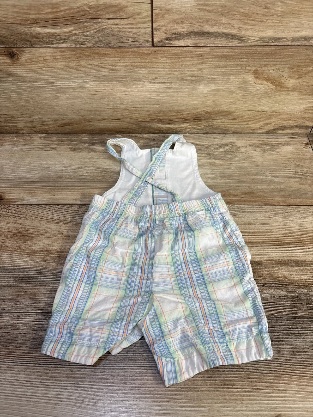 Back flat-lay of white baby plaid shortalls showing crossed straps and elastic waist with wood floor background