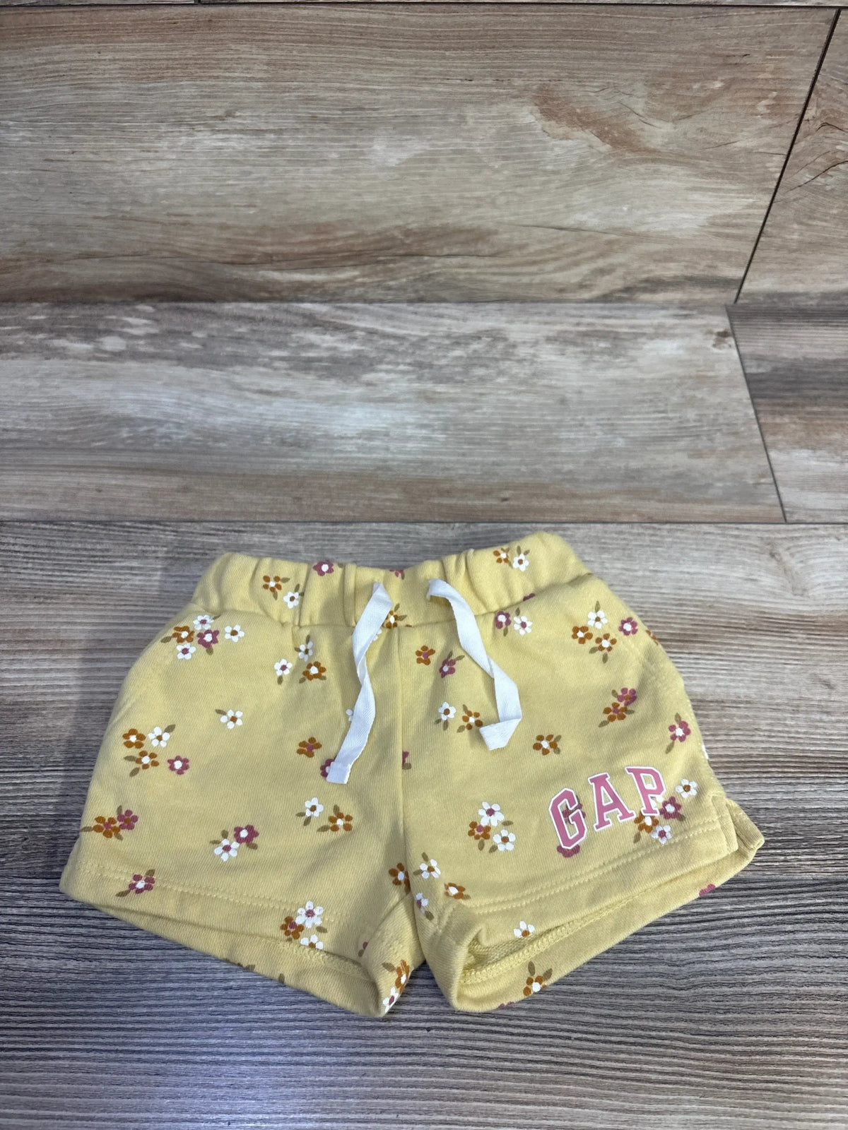 Flat lay of yellow Baby Gap shorts showing elastic waistband with white drawstring, small floral print, pink GAP logo on wood