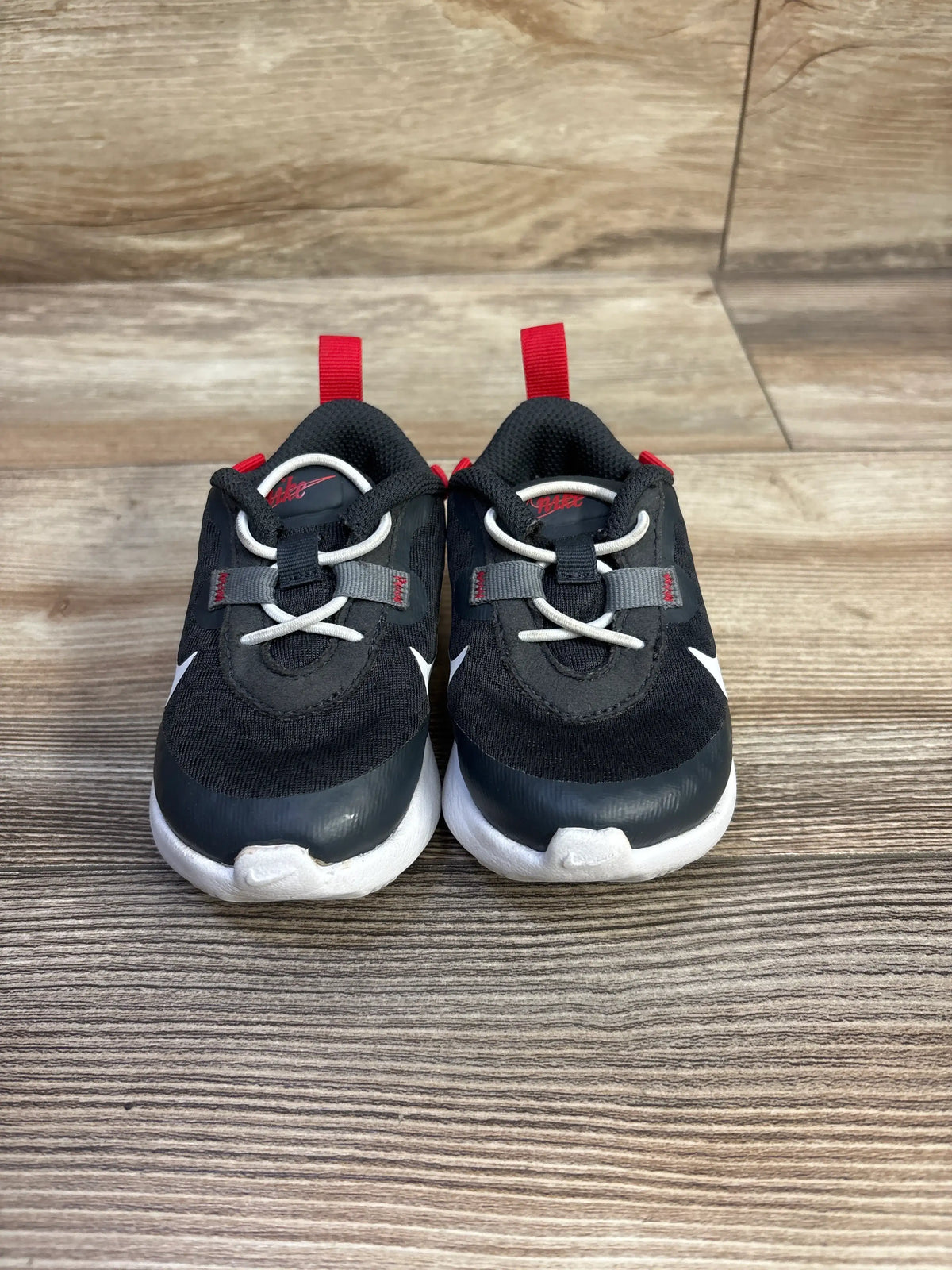 Front view of Nike Reposto TD Dark Smoke Grey sneakers showcasing red pull tabs and white soles on a woodgrain surface