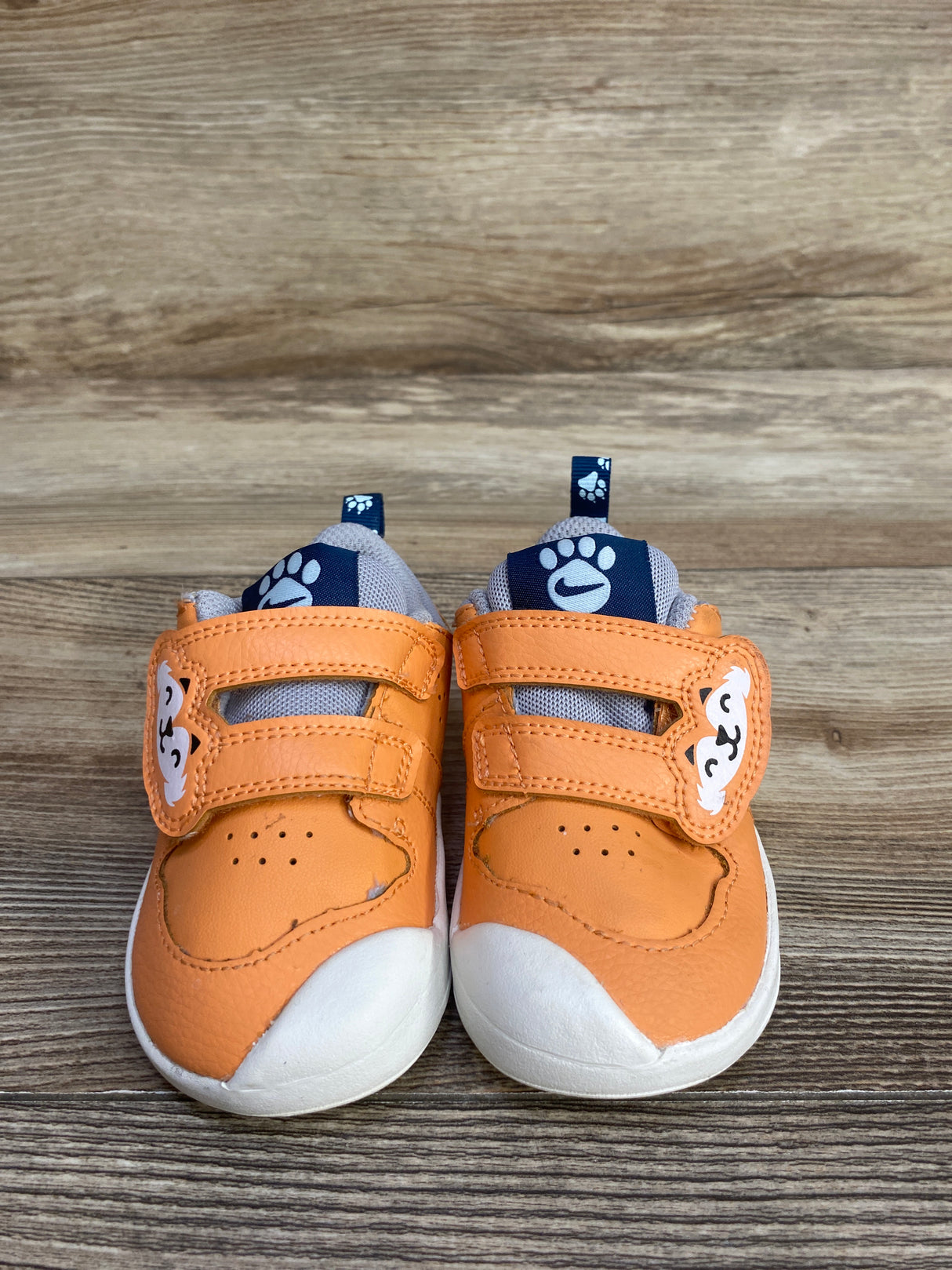 Pair of small orange toddler shoes with white soles, animal face details, and paw print tags on wooden floor