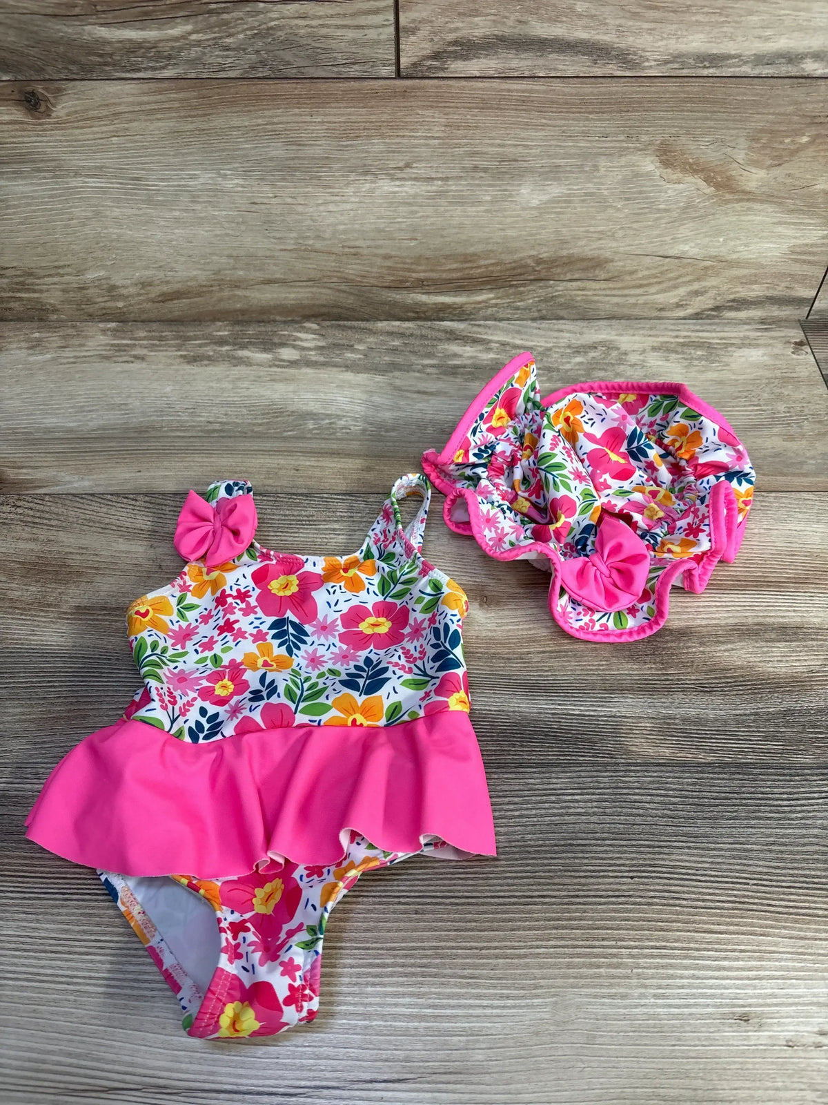 Flat lay of girls’ floral one-piece swimsuit and matching sunhat showcasing pink ruffle skirt, bow accents on wood background