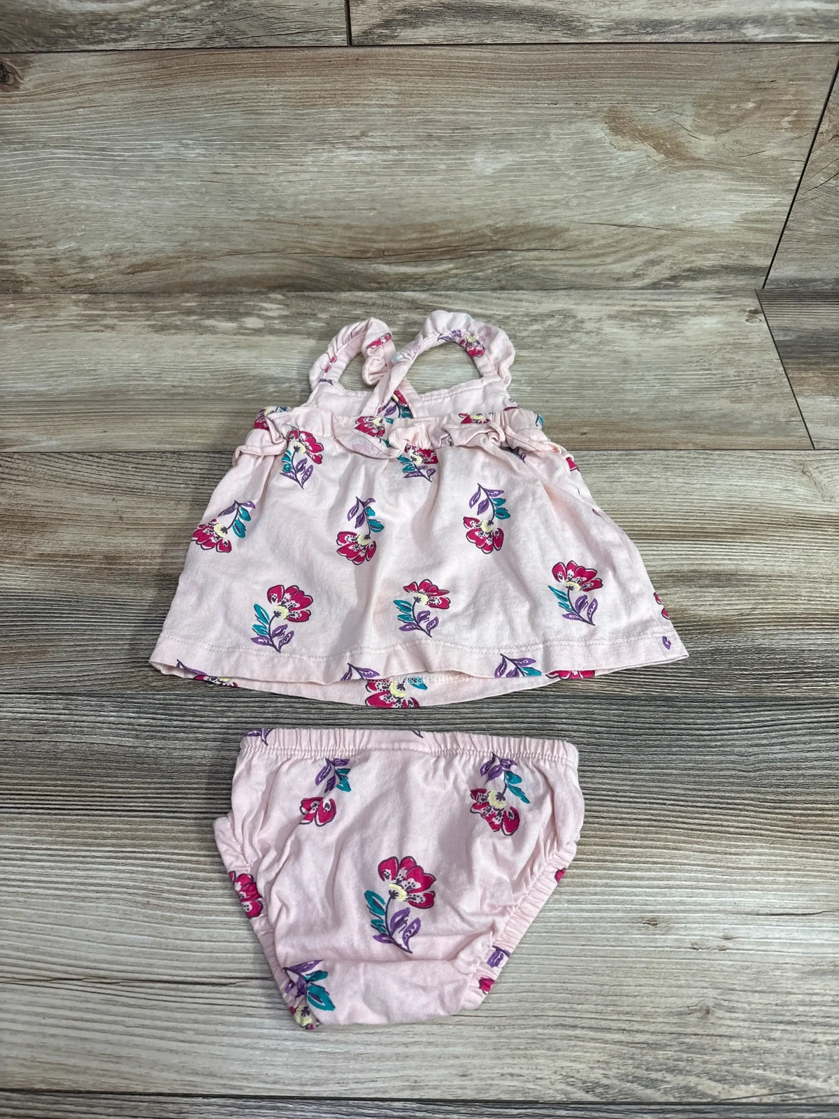 Flat lay of Carter’s pink 2-piece baby dress and bloomers showing ruffled straps and floral print on wood background