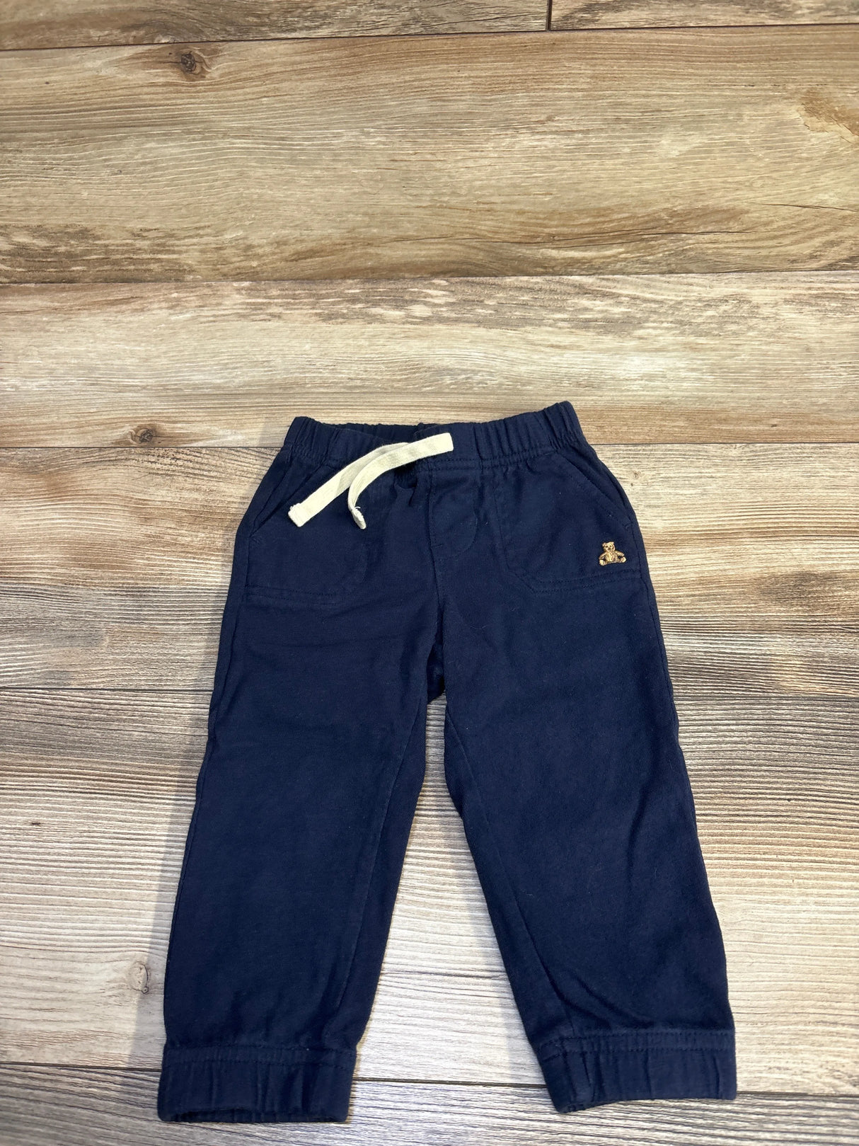 Top-down view of navy joggers with cream drawstring, front pockets, cuffed ankles and small embroidered bear on wood