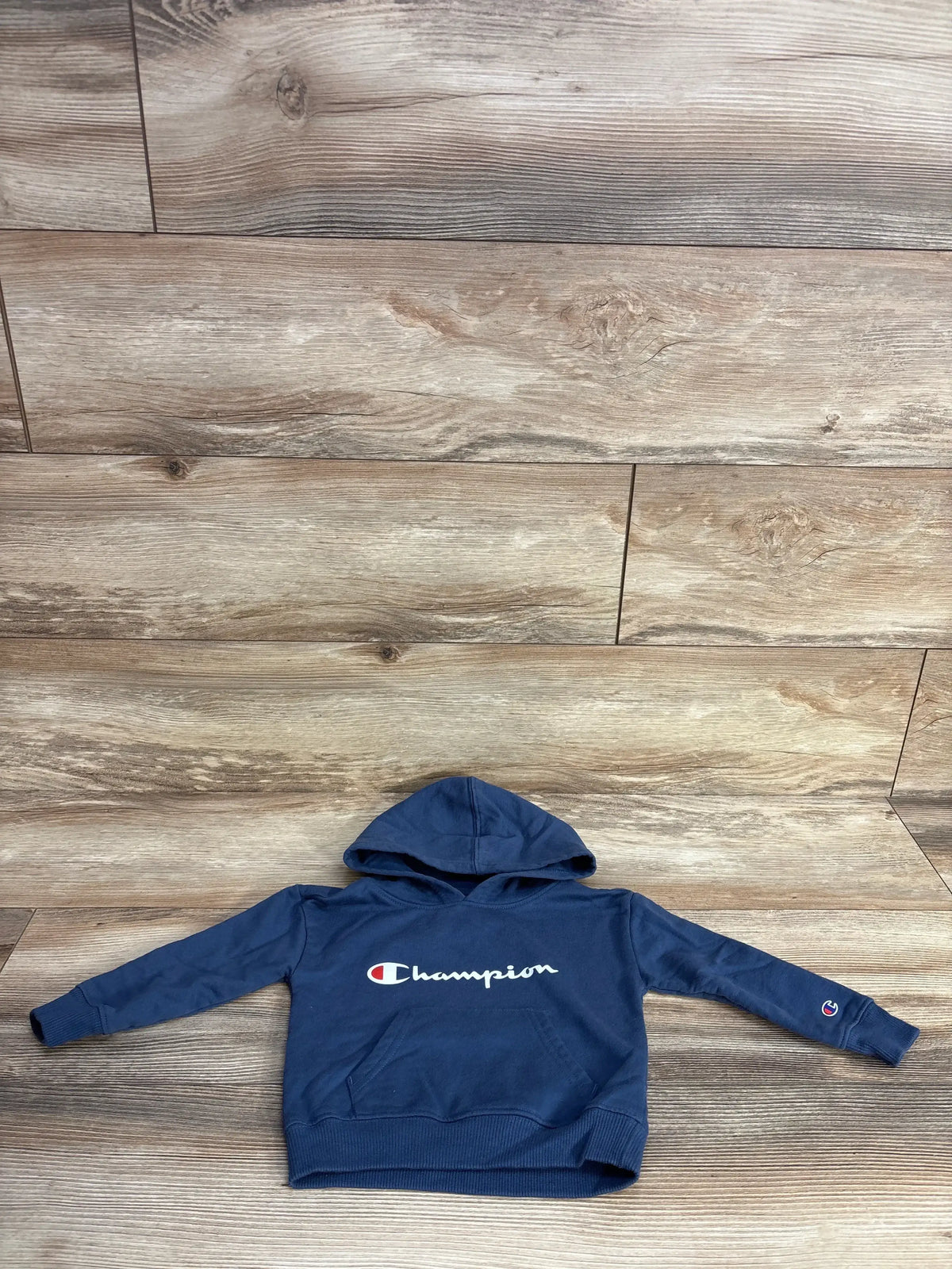 Navy blue Champion hoodie with front pocket and logo, laid flat on wood floor
