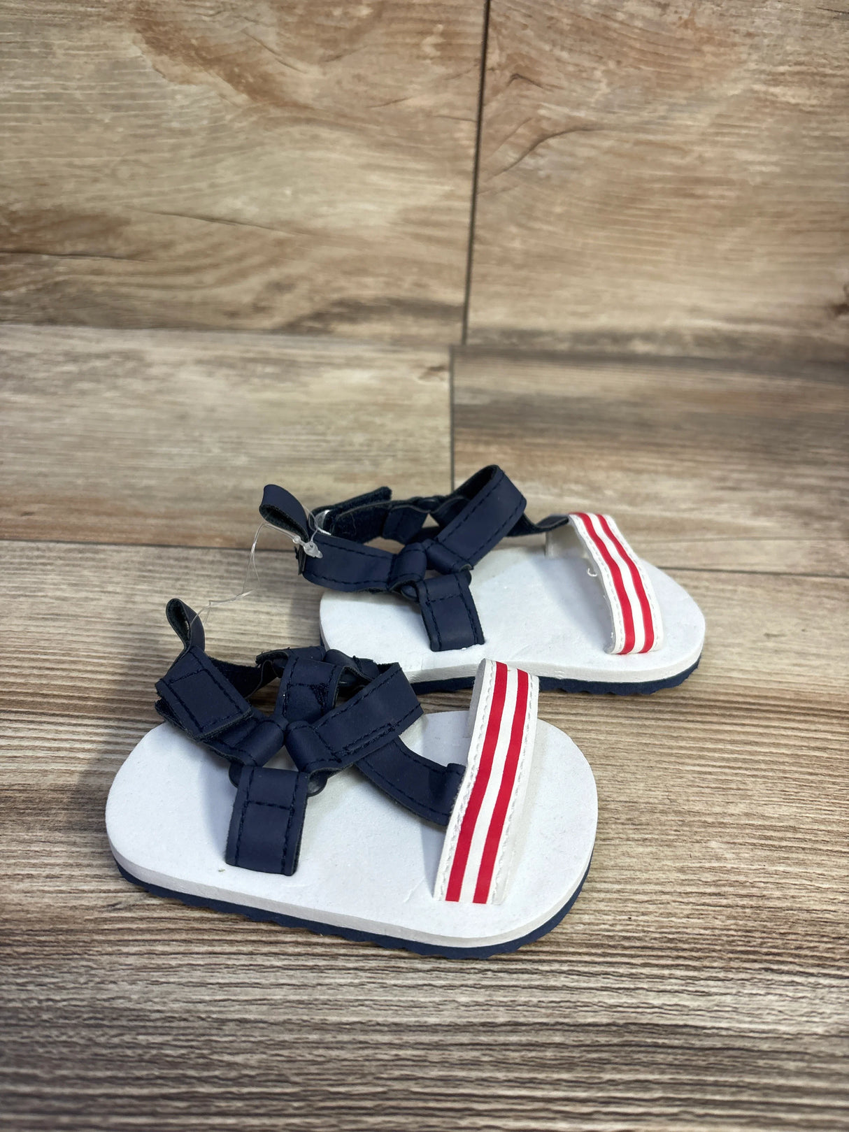 Front view of Child Of Mine baby sandals showcasing navy fabric straps and red and white striped toe band on wood floor background