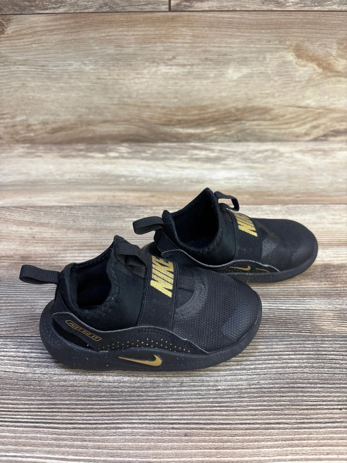 Angled side view of Nike Flex Runner TD toddler sneakers in black with gold NIKE strap, gold Swoosh and speckled midsole on wood floor, size 8C