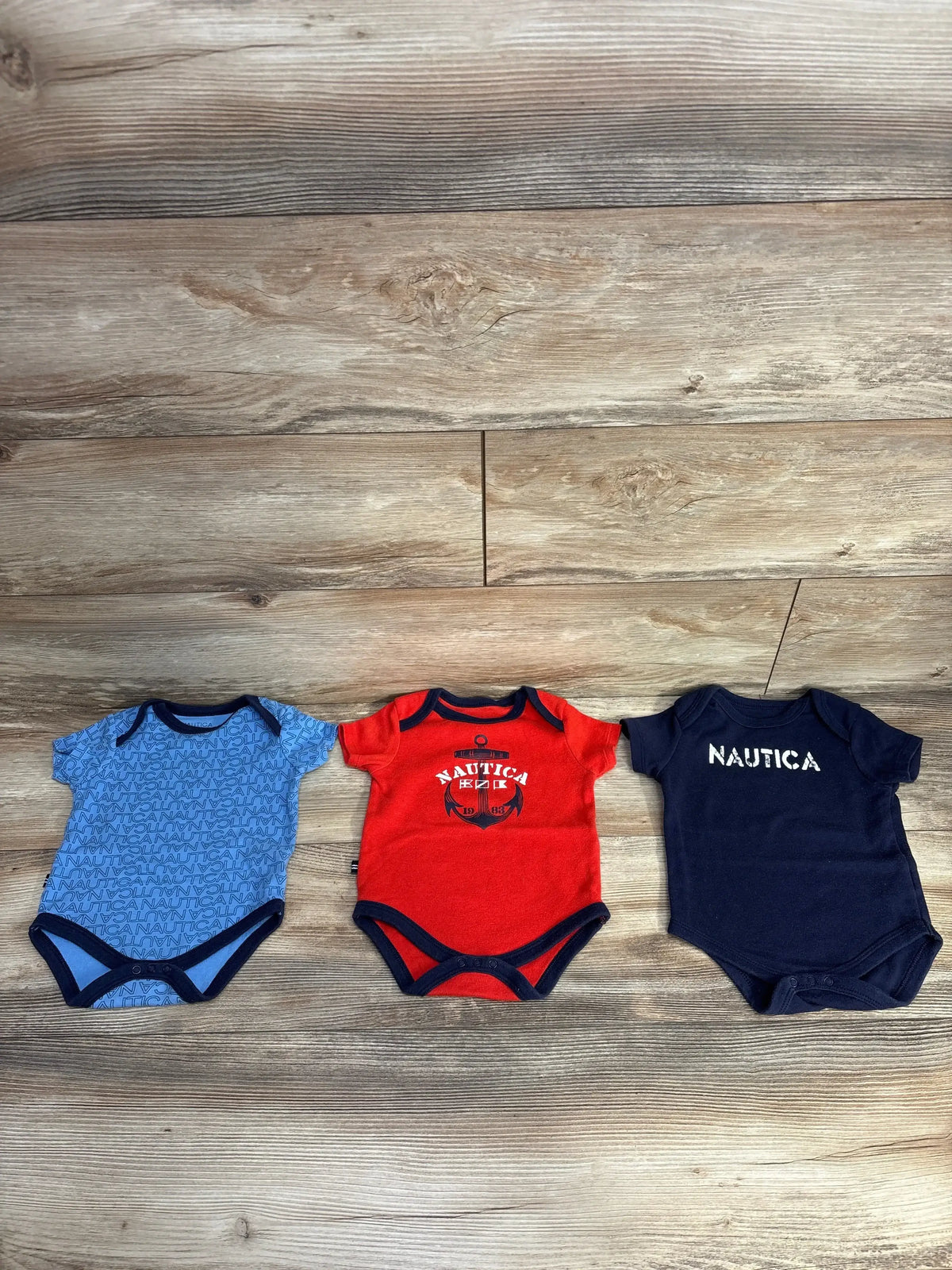 Three Nautica baby bodysuits in blue, red, and navy on wooden floor, kids clothing resale