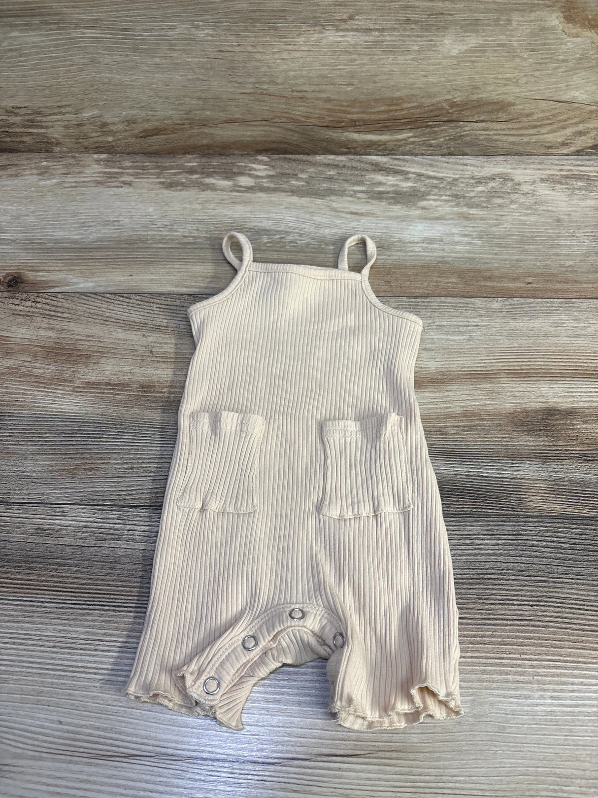 Flat lay of beige ribbed tank shortie romper (9-12 months) showcasing two front patch pockets and crotch snaps on wood background