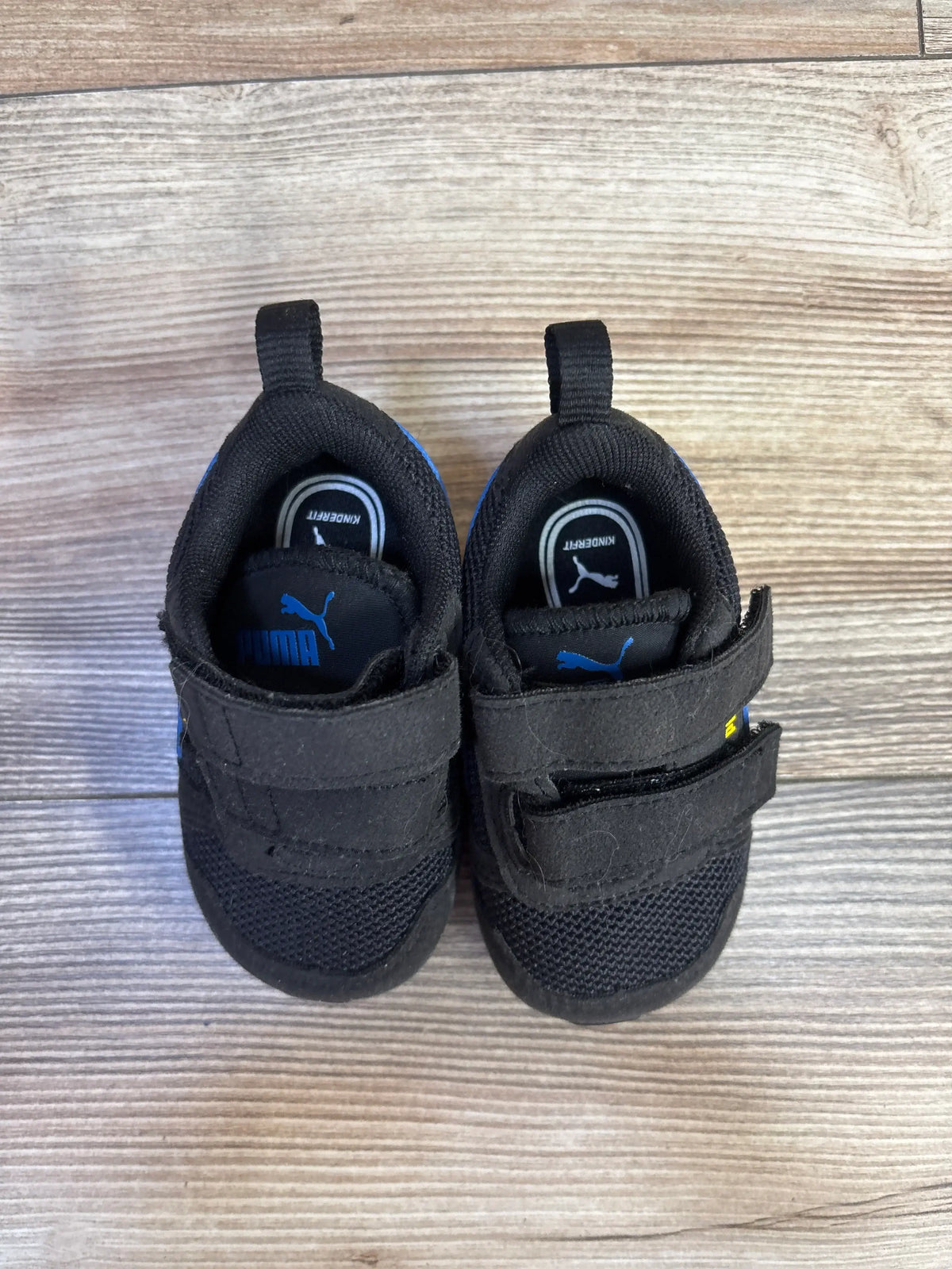 Top view of Puma R78 double strap sneakers showcasing black uppers and blue Puma logos with a wood-look background