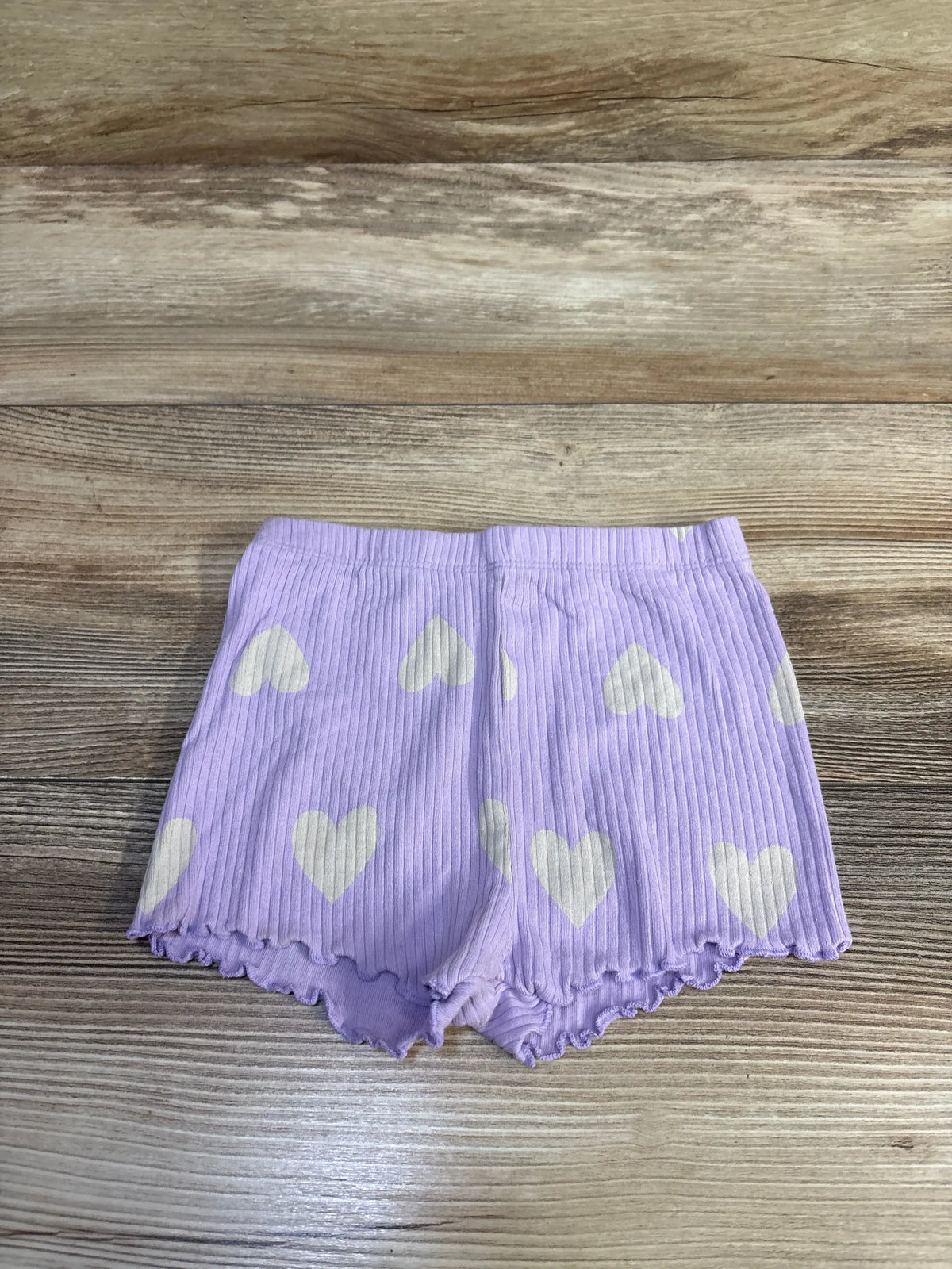 Front view of ribbed toddler bike shorts in lavender with white heart print and lettuce-edge hem on wood background