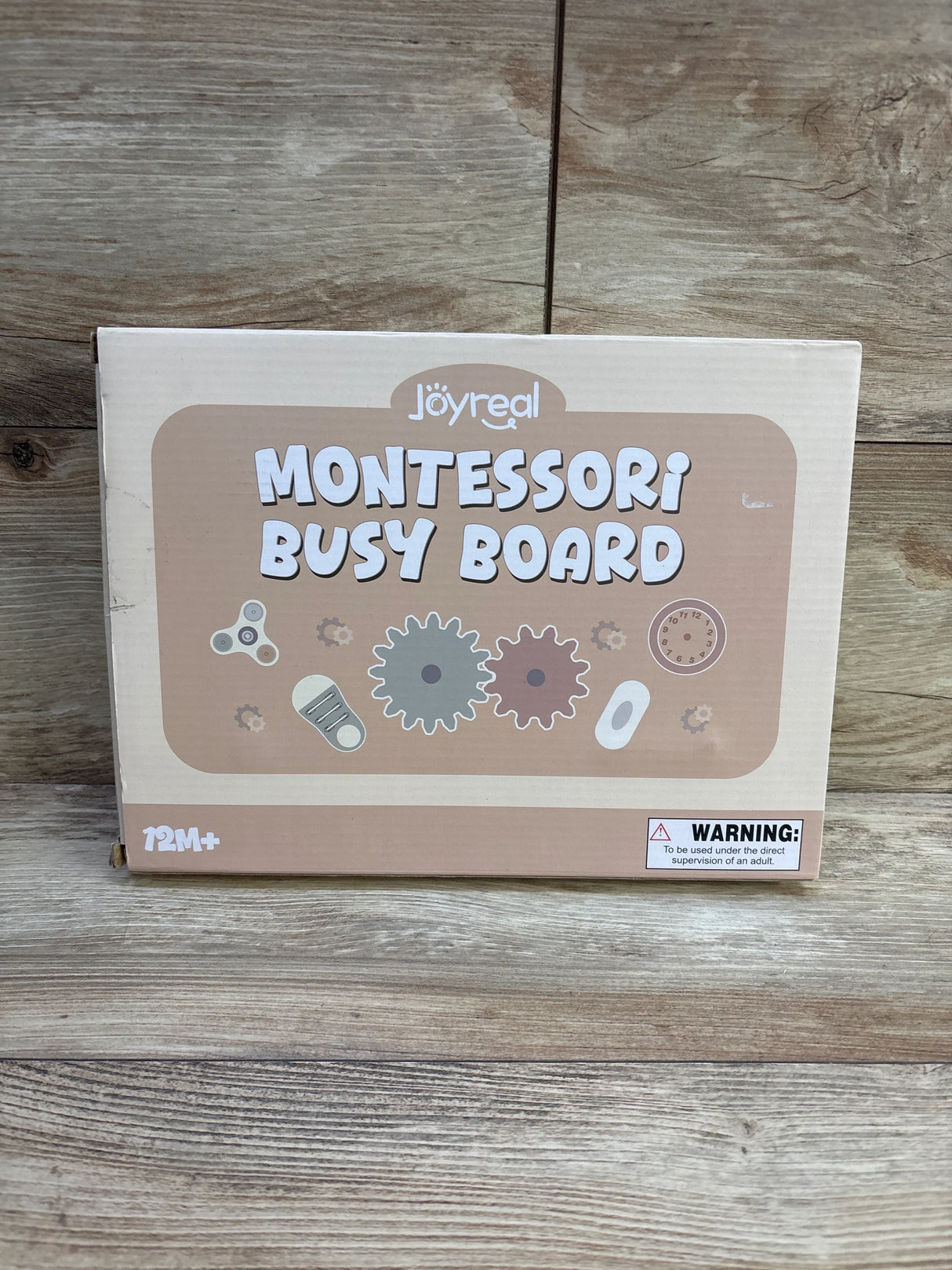 Front view of Joyreal busy board box showing MONTESSORI BUSY BOARD text, gear icons and warning label on wood background