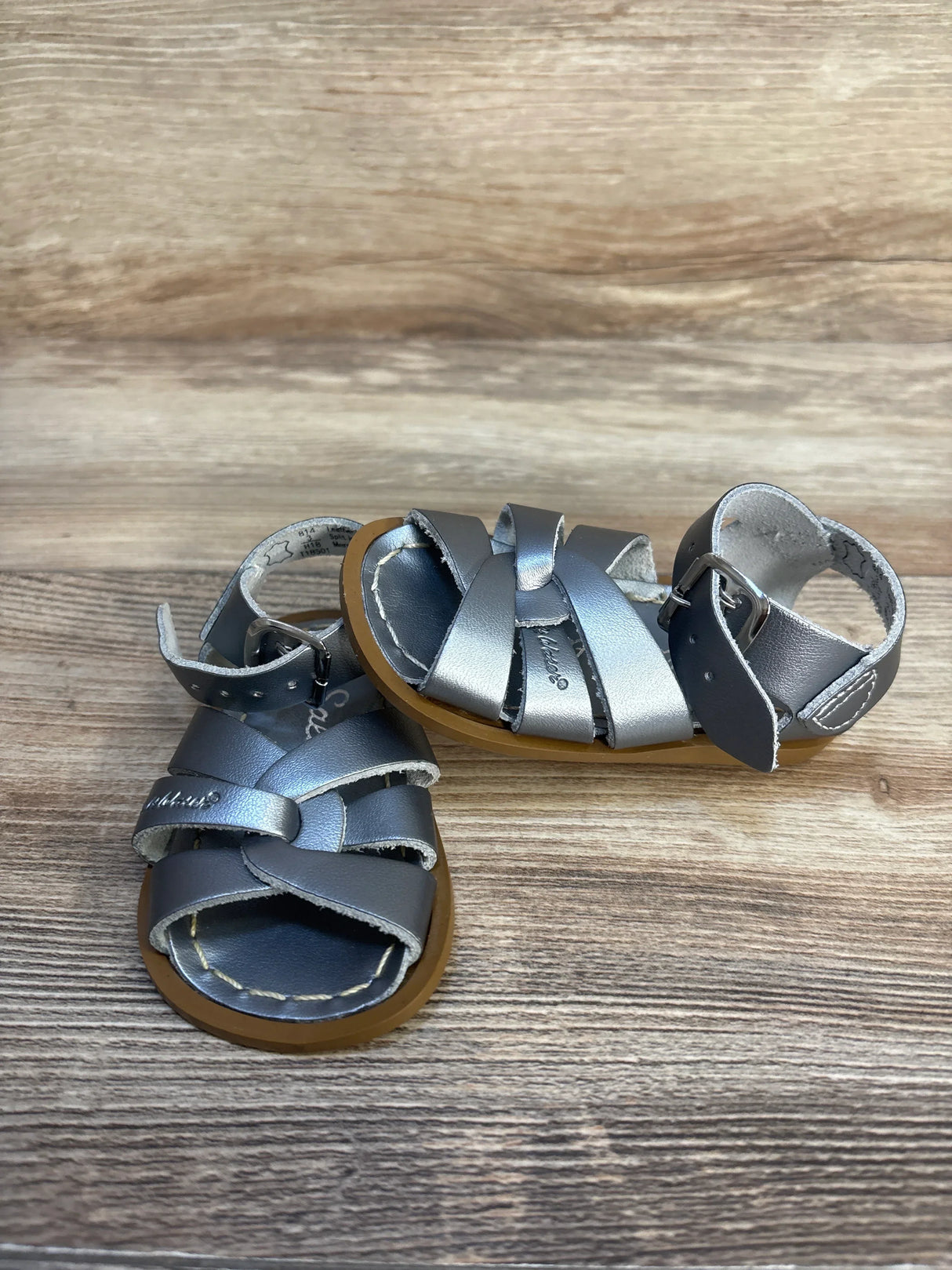 Silver toddler sandals with crisscross leather straps and brown soles on wood background
