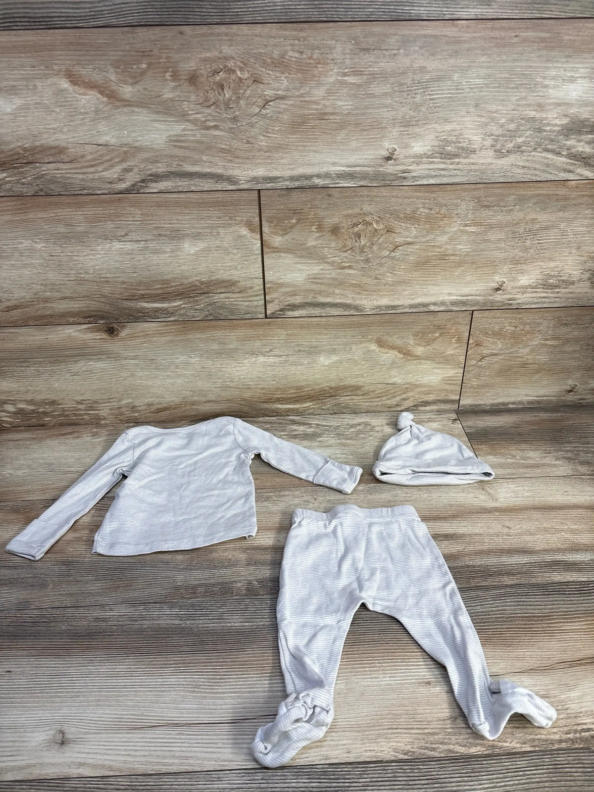 Flat lay of 3-piece beige striped baby set: long-sleeve shirt, footed pants, and knot-top hat, on wood plank background