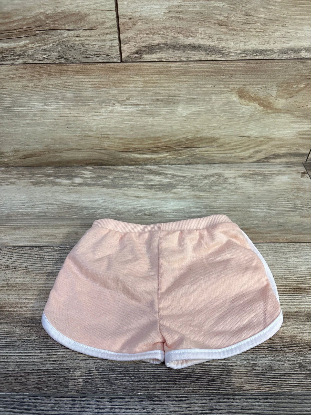 Top-down view of Just Lizzy toddler shorts, pink with elastic waistband and white side trim, size 12 months, laid flat on wood background