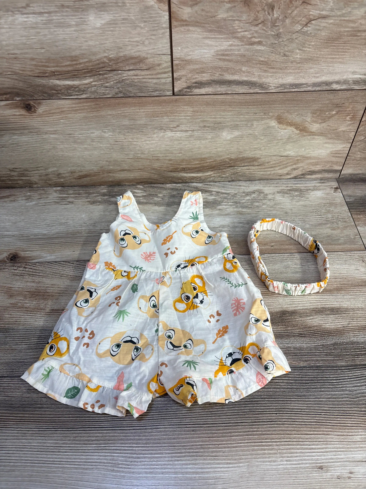 Front view of Disney Baby The Lion King romper and headband set showcasing an allover character print on a wood background