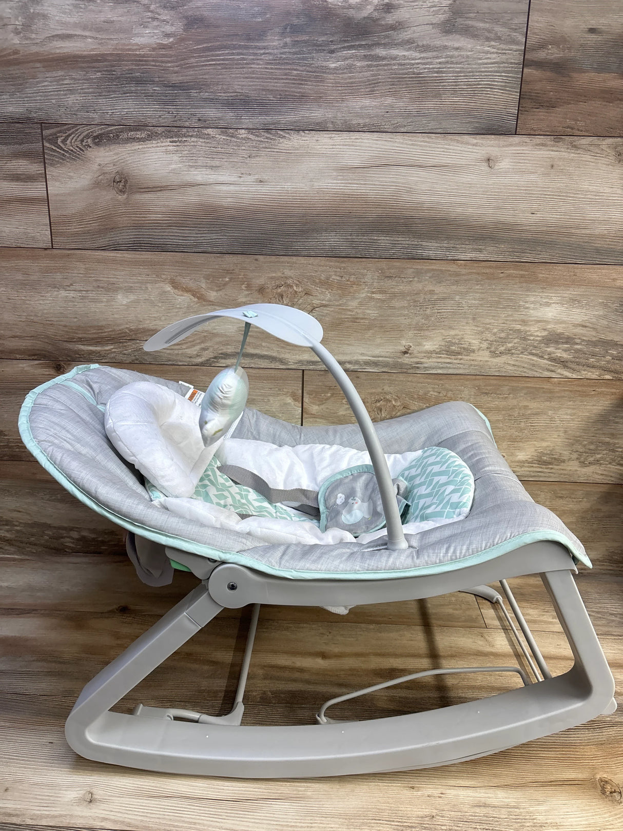 Side view of Ingenuity Keep Cozy 3-in-1 baby bouncer showing gray and mint padded seat with toy bar on wood floor