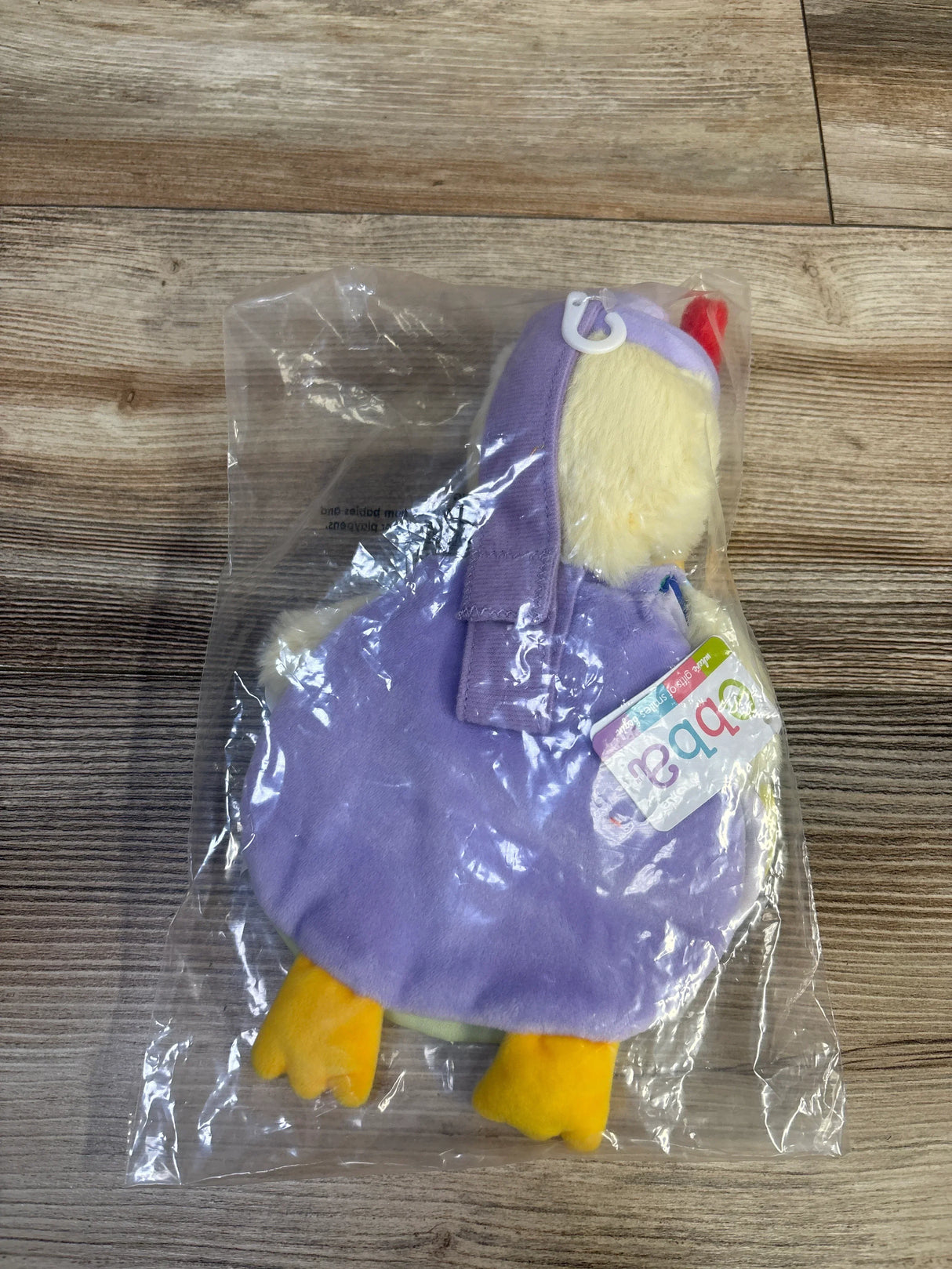 Top-down view of Mother Goose plush book showing purple cover, yellow plush feet and tag, in clear plastic bag on wood background