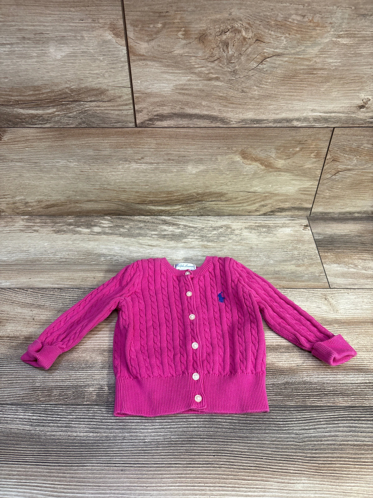Front view of a pink Ralph Lauren mini cable cardigan showcasing cable knit texture and button front with a wood floor background