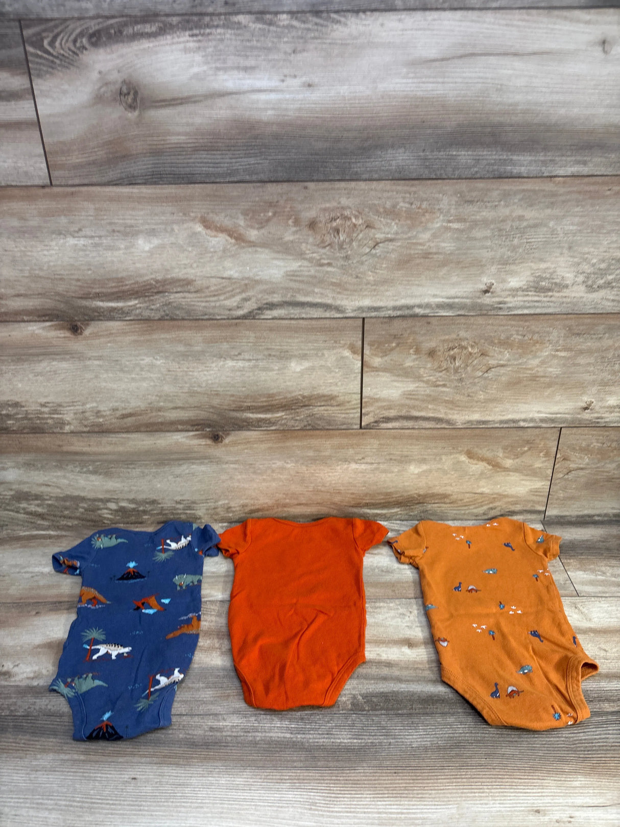 Top-down view of three Carter’s baby bodysuits showcasing a blue dinosaur print, a solid orange, and an orange animal print on a wood plank background