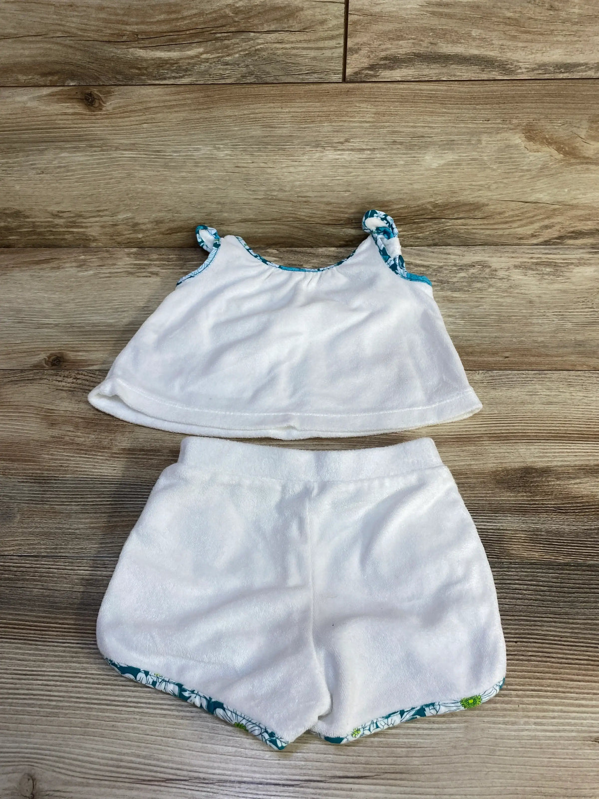 Flat lay of Janie and Jack two-piece terry set in white showcasing tank top and shorts with blue green floral trim on wood plank floor