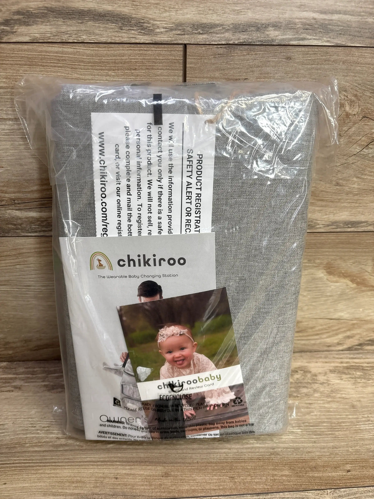 Front view of new Chikiroo wearable baby changing station in gray sealed packaging on wood plank floor