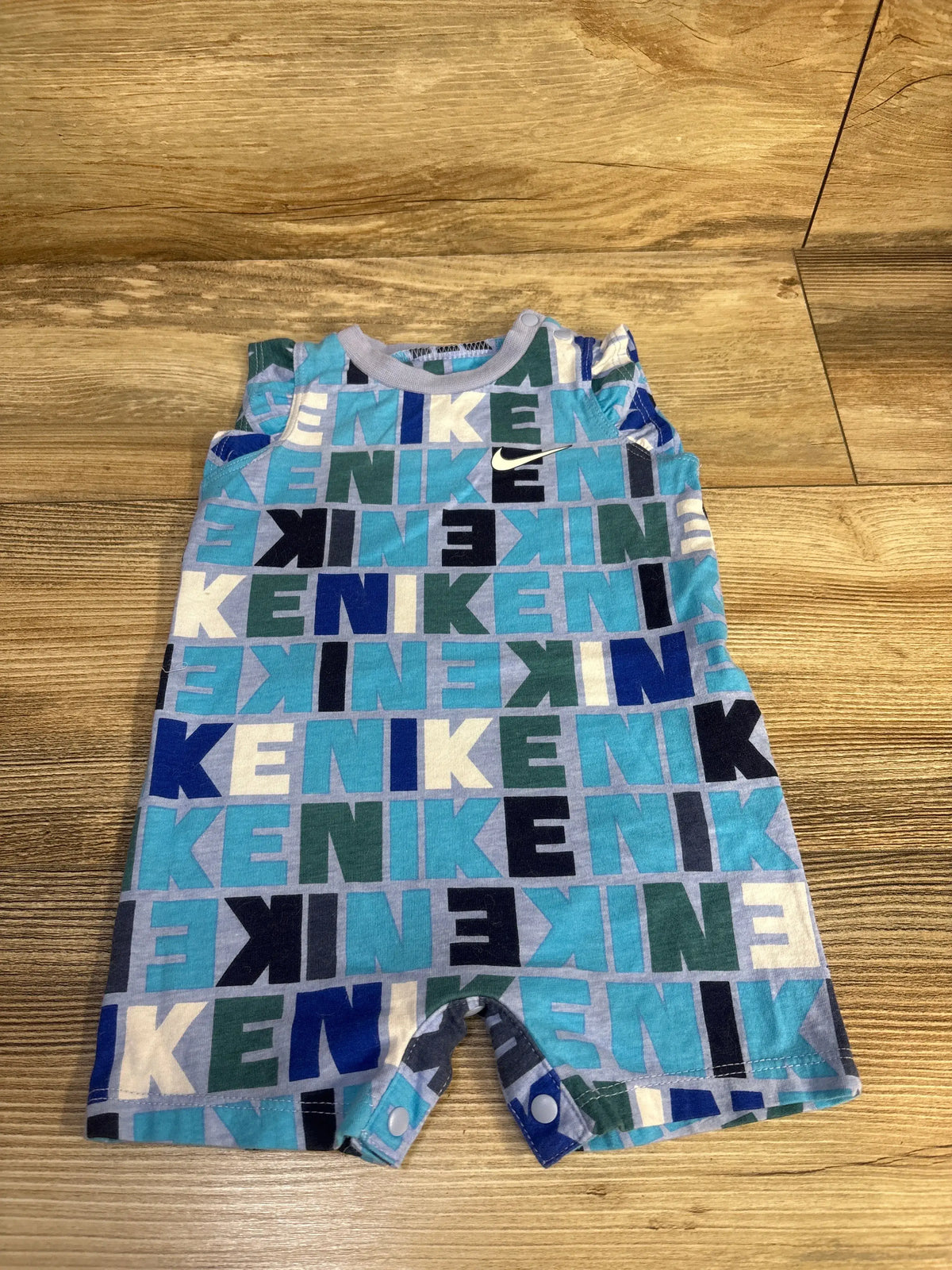 Front view of Nike blue flutter sleeve romper showcasing repeating Nike lettering and snap closures on a wood floor