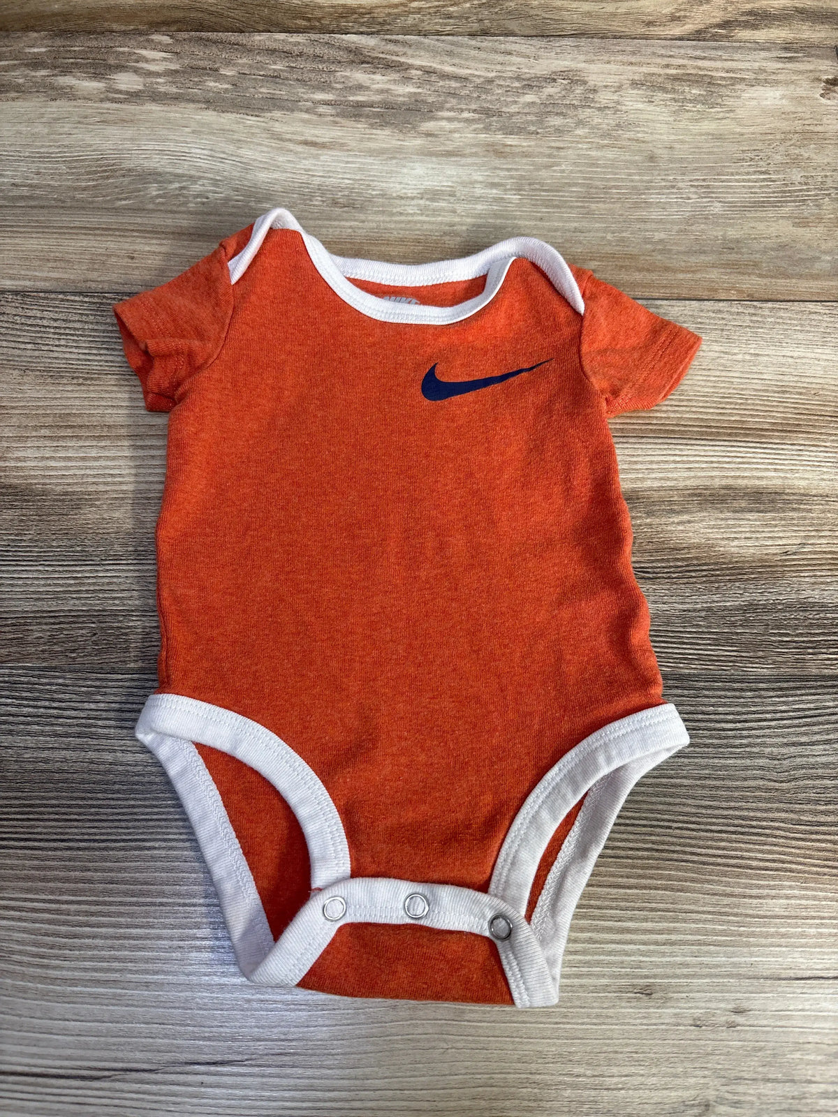 Front view of Nike orange bodysuit showcasing white trim, short sleeves, Nike swoosh, and snap closures on a wood-grain background