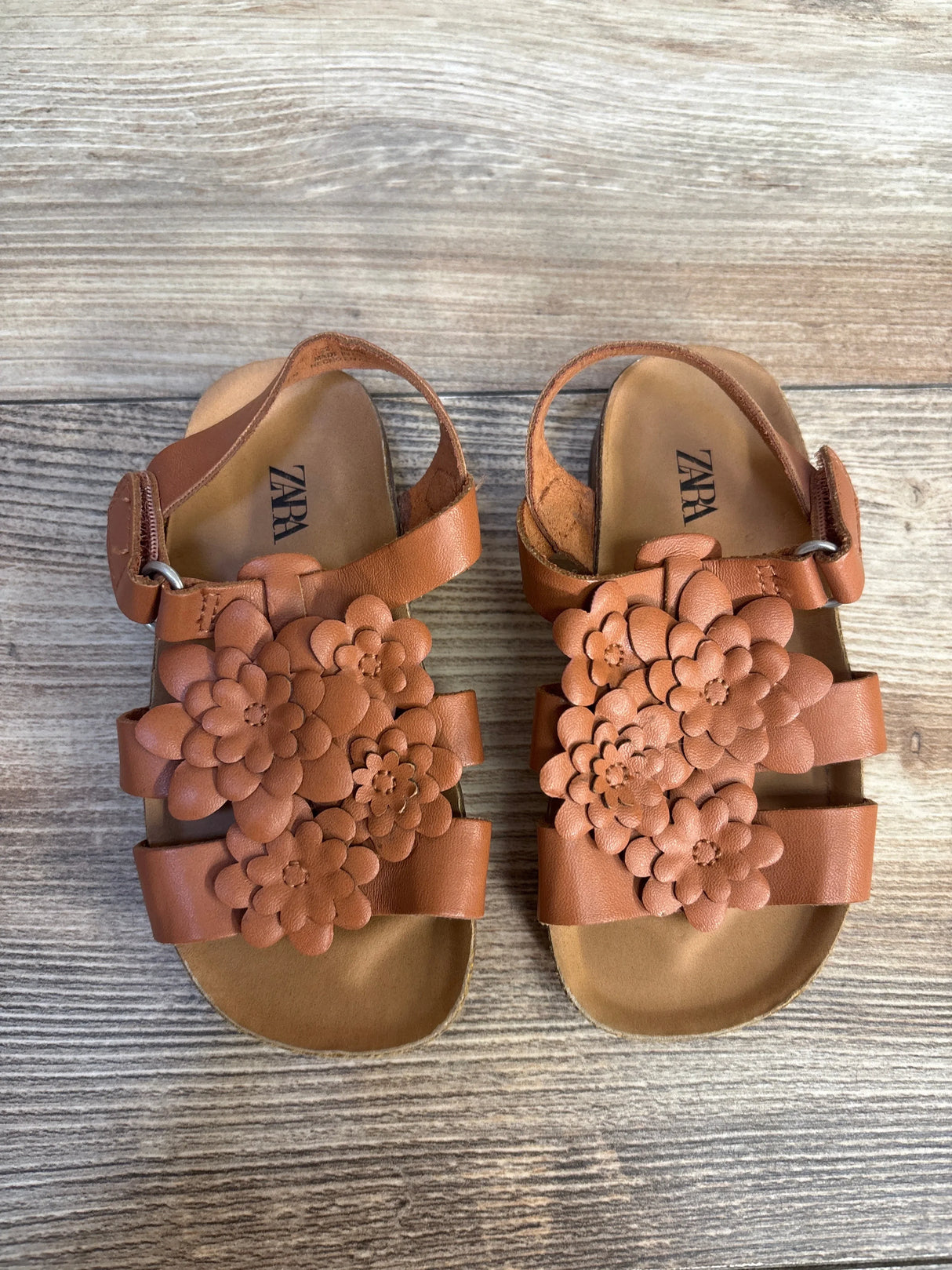 Brown toddler sandals with floral accents and ankle straps on wooden background