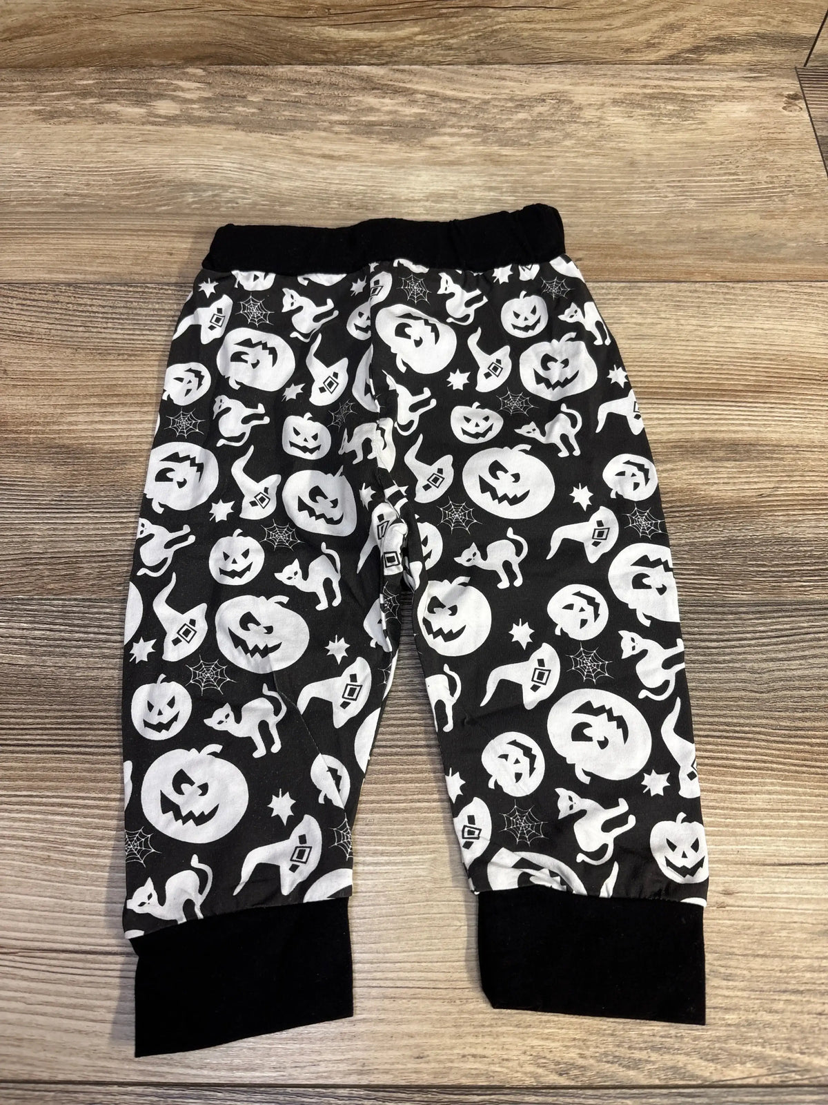 Front view of black Halloween pants showcasing pumpkin, ghost, cat, witch hat, and spiderweb pattern on a wood background