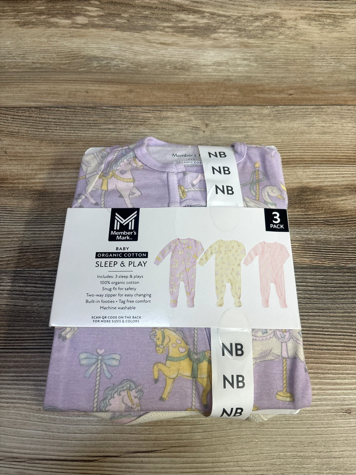 Front view of folded Member's Mark 3-pack newborn organic cotton sleep-and-play set in lavender carousel print with packaging label showing NB, on wood surface