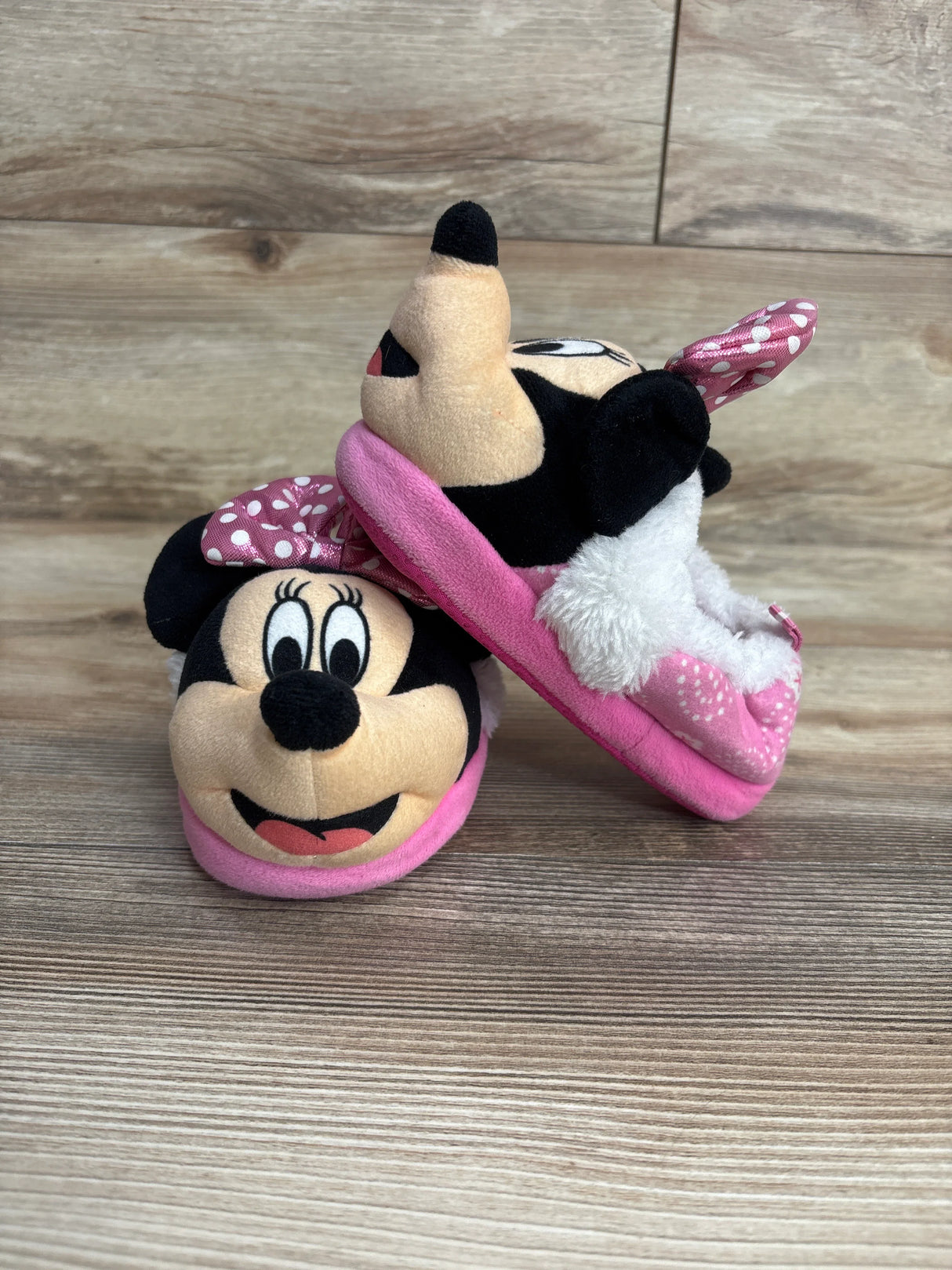 Pair of pink Minnie Mouse plush slippers for kids on wooden floor, featuring polka dot bow and fuzzy lining