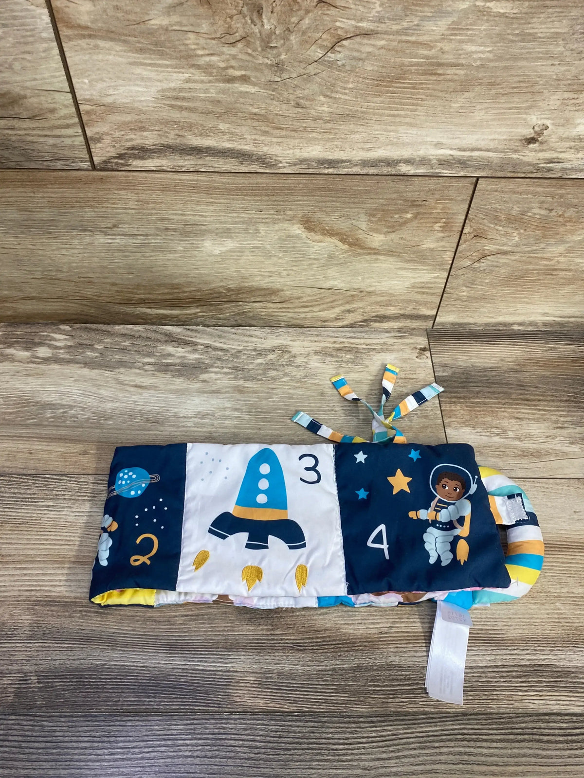 Front view of Little Muffincakes soft baby book showing rocket and astronaut panels with numbers, fabric ring and ribbon tags on wood floor