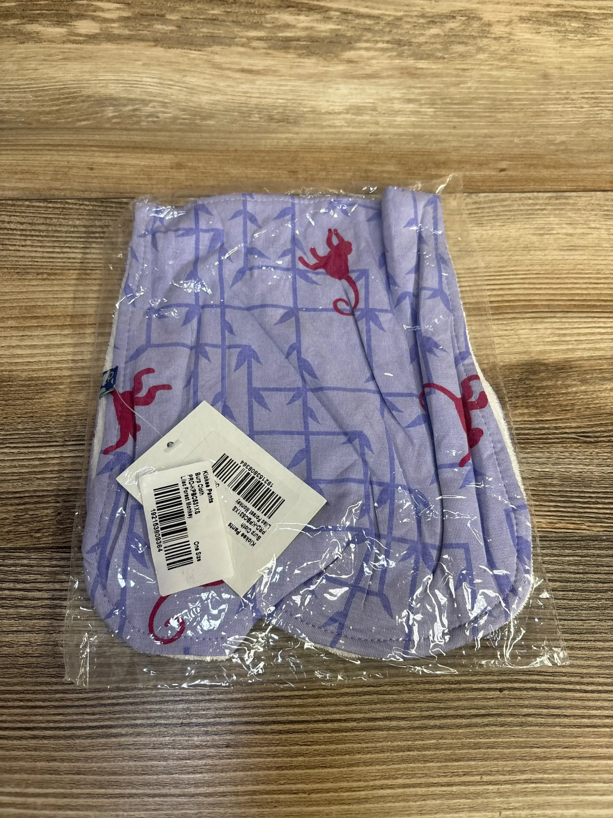 Folded Kickee Pants burp cloth in lilac showing purple bamboo-grid and red monkey print in clear plastic on wood surface