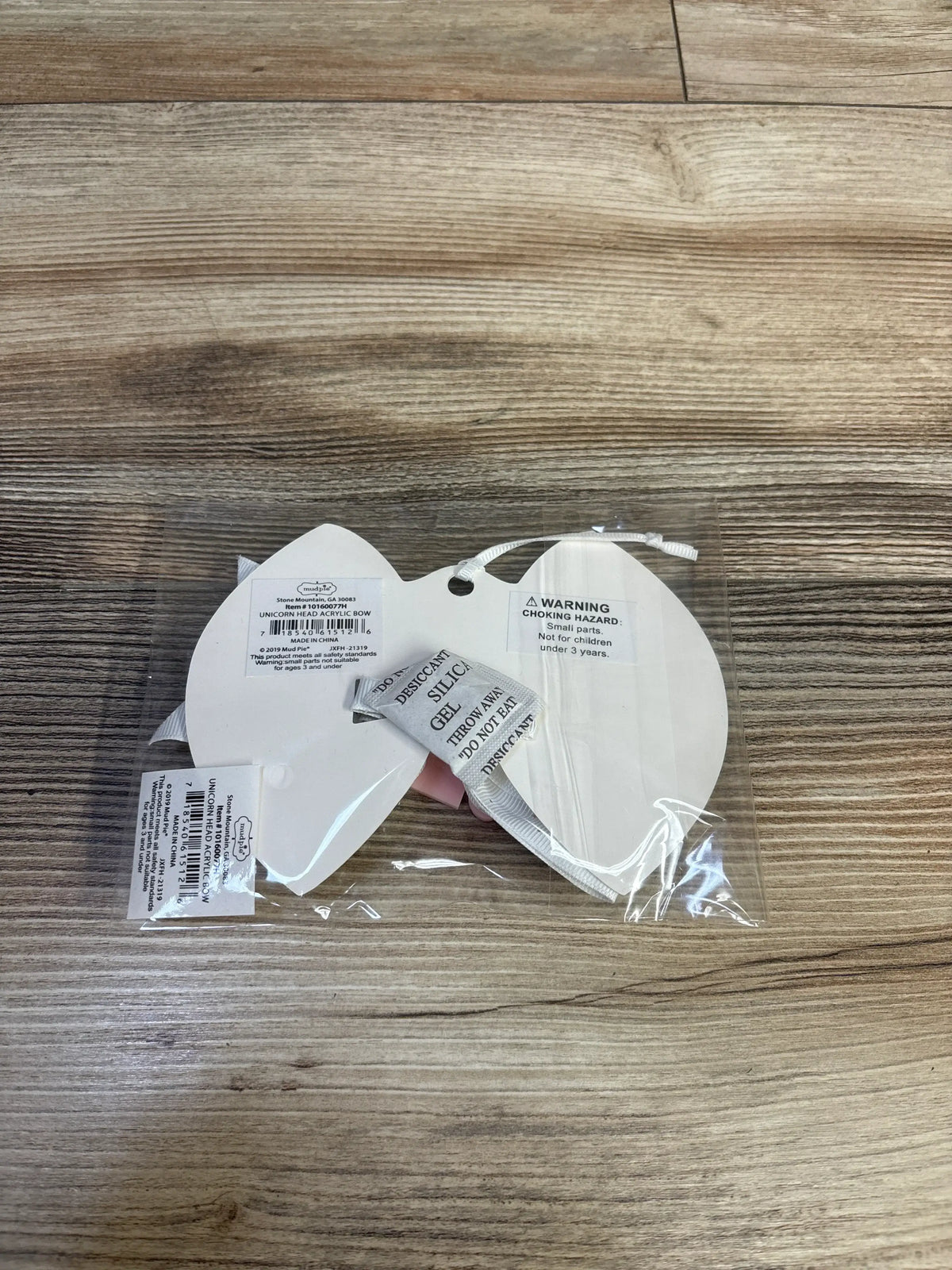 Back view of white Mud Pie unicorn head acrylic bow in clear packaging on a wood-grain background