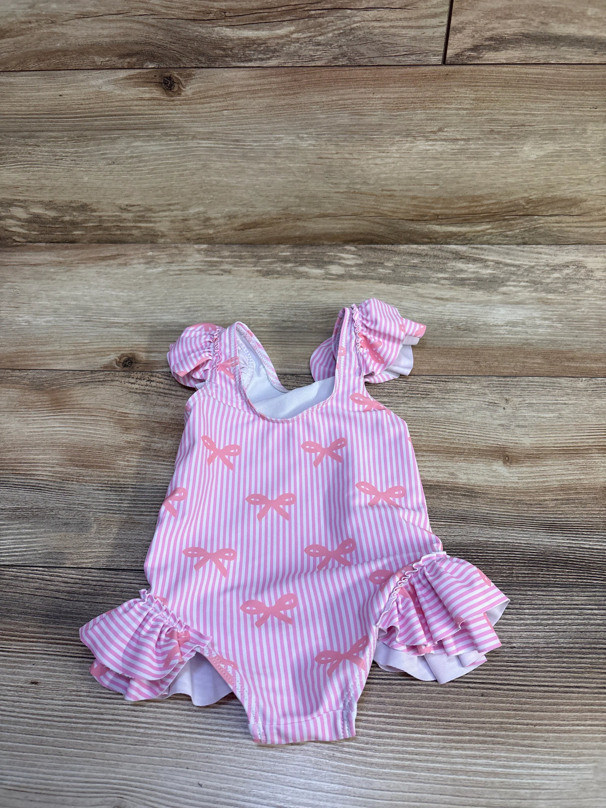 Front view of pink striped baby one-piece swimsuit with bow print and ruffled shoulder and leg trim on wood background