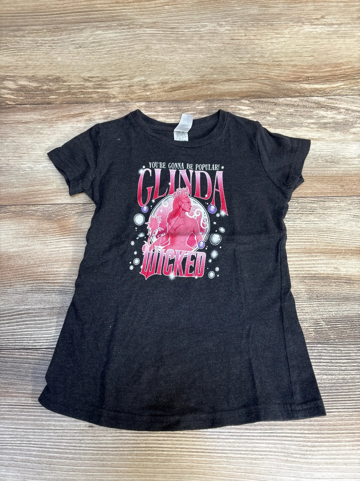 Black kids t-shirt with Glinda Wicked graphic and text, laid flat on wood background