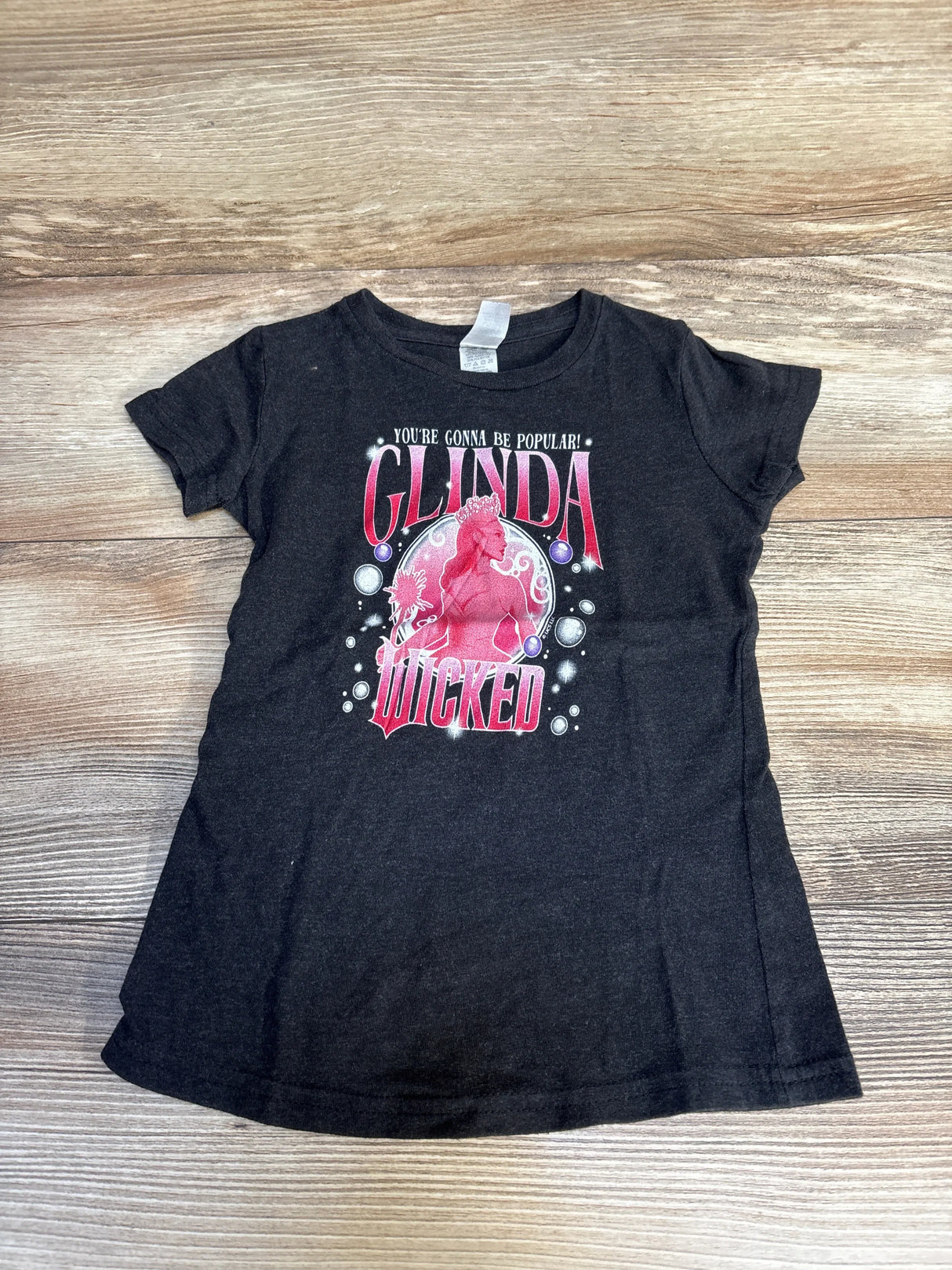Black kids t-shirt with Glinda Wicked graphic and text, laid flat on wood background